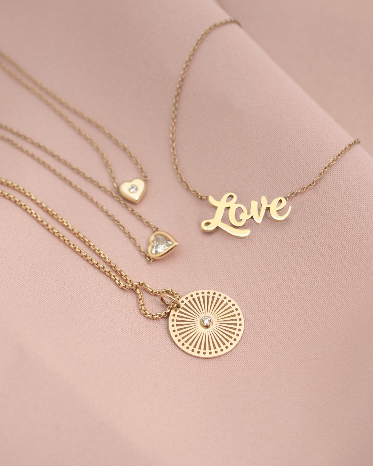 Four Zoë Chicco 14k gold and diamond Valentine's Day collection necklaces laying flat against a light pink background