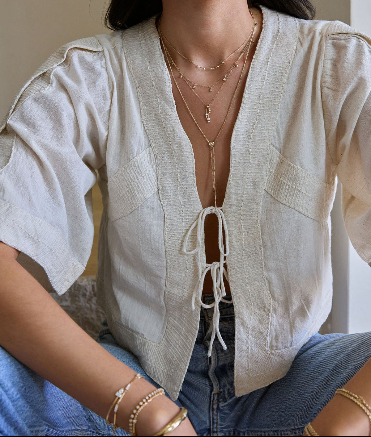 Person wearing a light-colored linen cardigan with layers of Zoe Chicco 14k gold and diamond necklace and bracelets, sitting indoors.