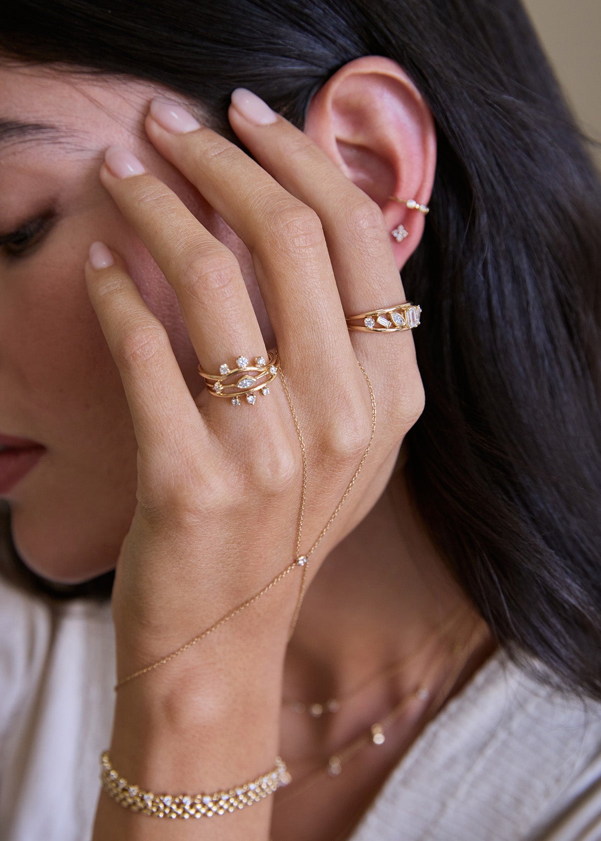 Close-up of a woman's hand and ear with Zoe Chicco 14k gold and diamond rings and hand chain on a neutral background
