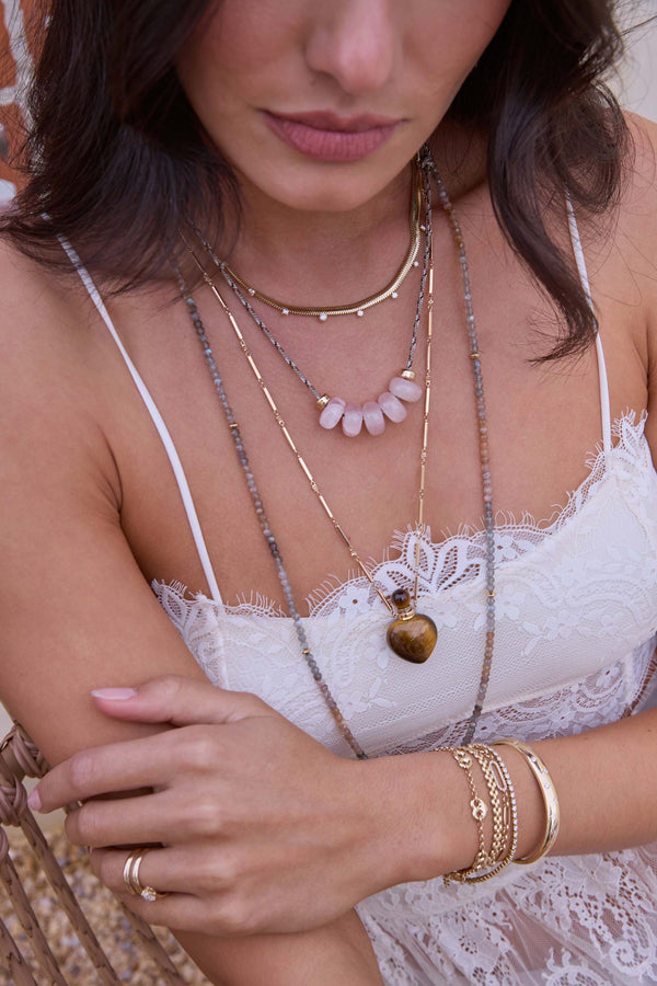 Close-up of a woman wearing Zoe Chicco 14k gold necklaces and bracelets with paracord, rose quartz beads, and a tiger eye perfume bottle with a blurred background