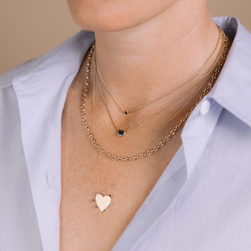 woman in a light blue shirt wearing a Zoë Chicco 14k Gold Blue Sapphire Bezel XS Curb Chain Necklace layered with a 14k XS Curb Chain Emerald Cut Blue Sapphire Bezel Necklace, Small Puffed Mariner Chain Necklace, and a 14k 7 Rainbow Gemstones Heart Pendant Necklace