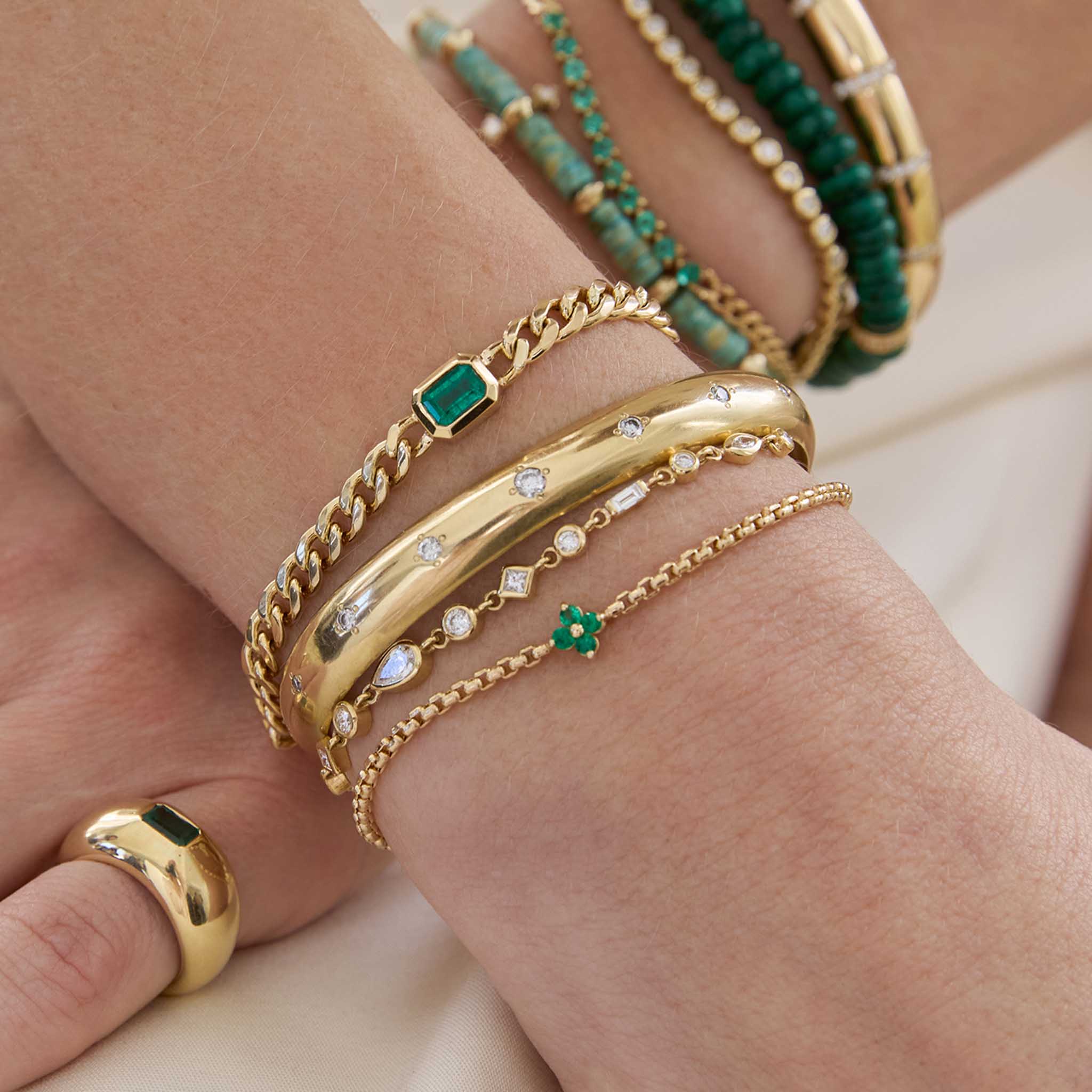 woman's wrist wearing a Zoë Chicco 14k Gold Prong Emerald Quad Box Chain Bracelet stacked with a 14k Large Linked Mixed Cut Diamond Bolo Bracelet, 14k Graduated Diamonds Wide Half Round Cuff, and a 14k Medium Curb Chain Emerald Cut Emerald Bezel Bracelet