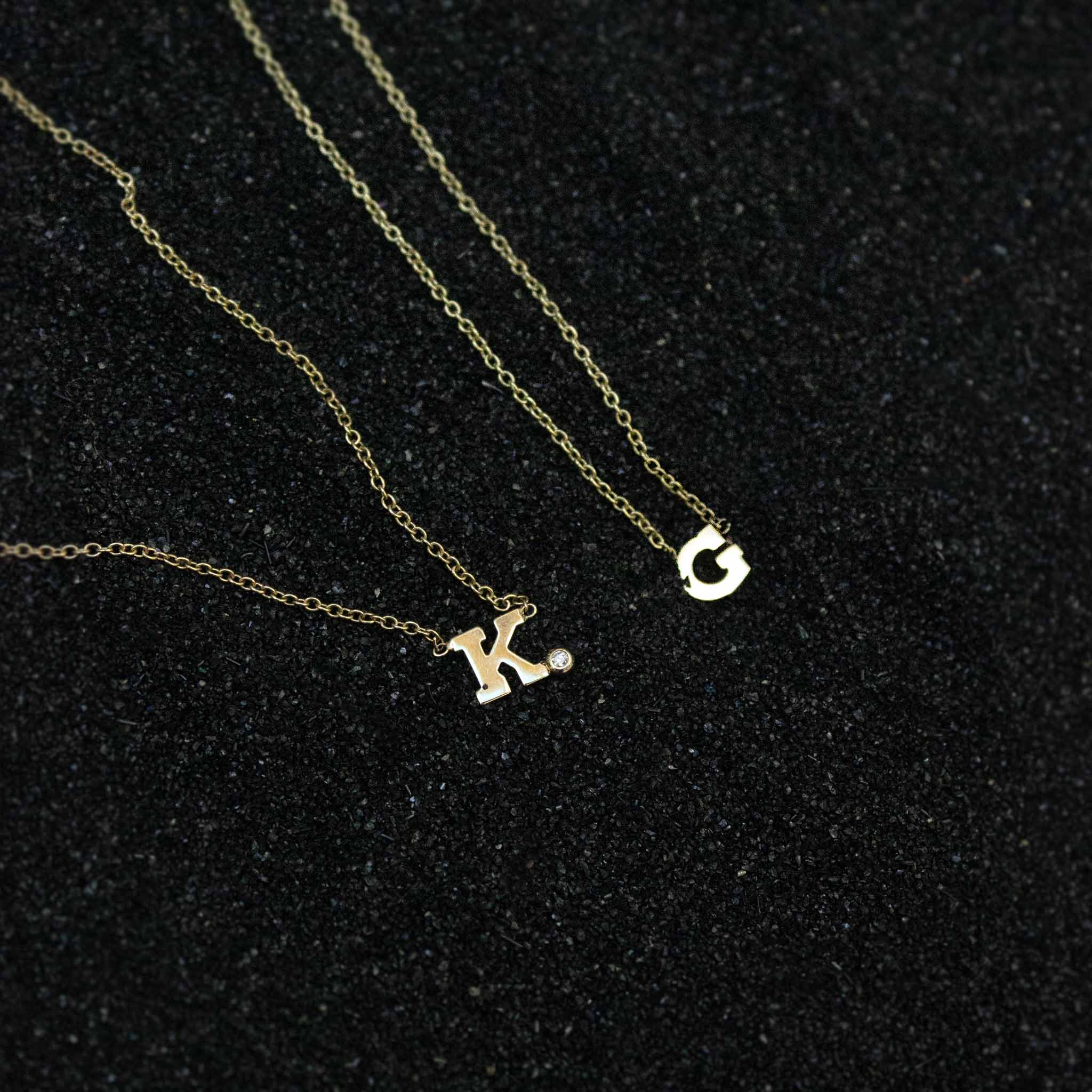 On black sand lies two necklaces: a letter necklace with bezel set diamond shown as "K" and a plain 14k gold block letter shown as "G".