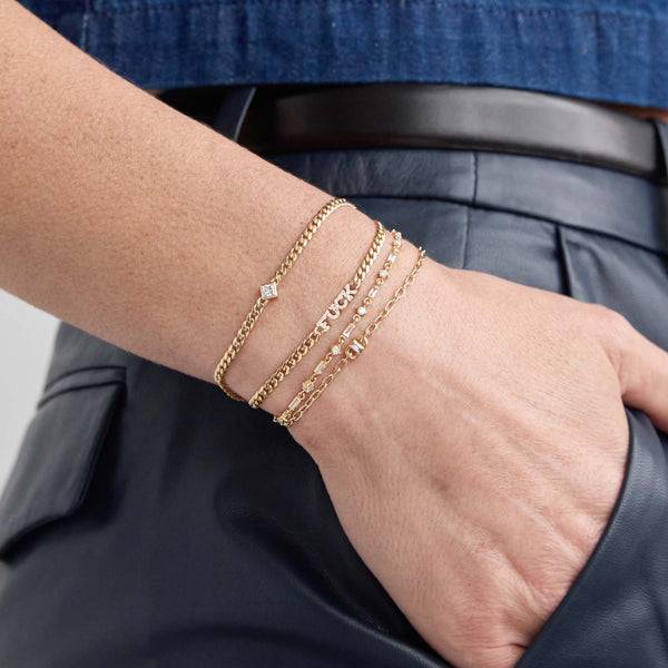 Woman with her hands in her right pocket wearing a Zoë Chicco 14k Gold Baguette Diamond Open Link Square Oval Chain Bracelet on her wrist layered with a 14k Linked Baguette & Prong Diamond Tennis Bracelet, 14k Princess Diamond Small Curb Chain Bracelet, and a 14k Itty Bitty Pavé Diamond FUCK Bracelet