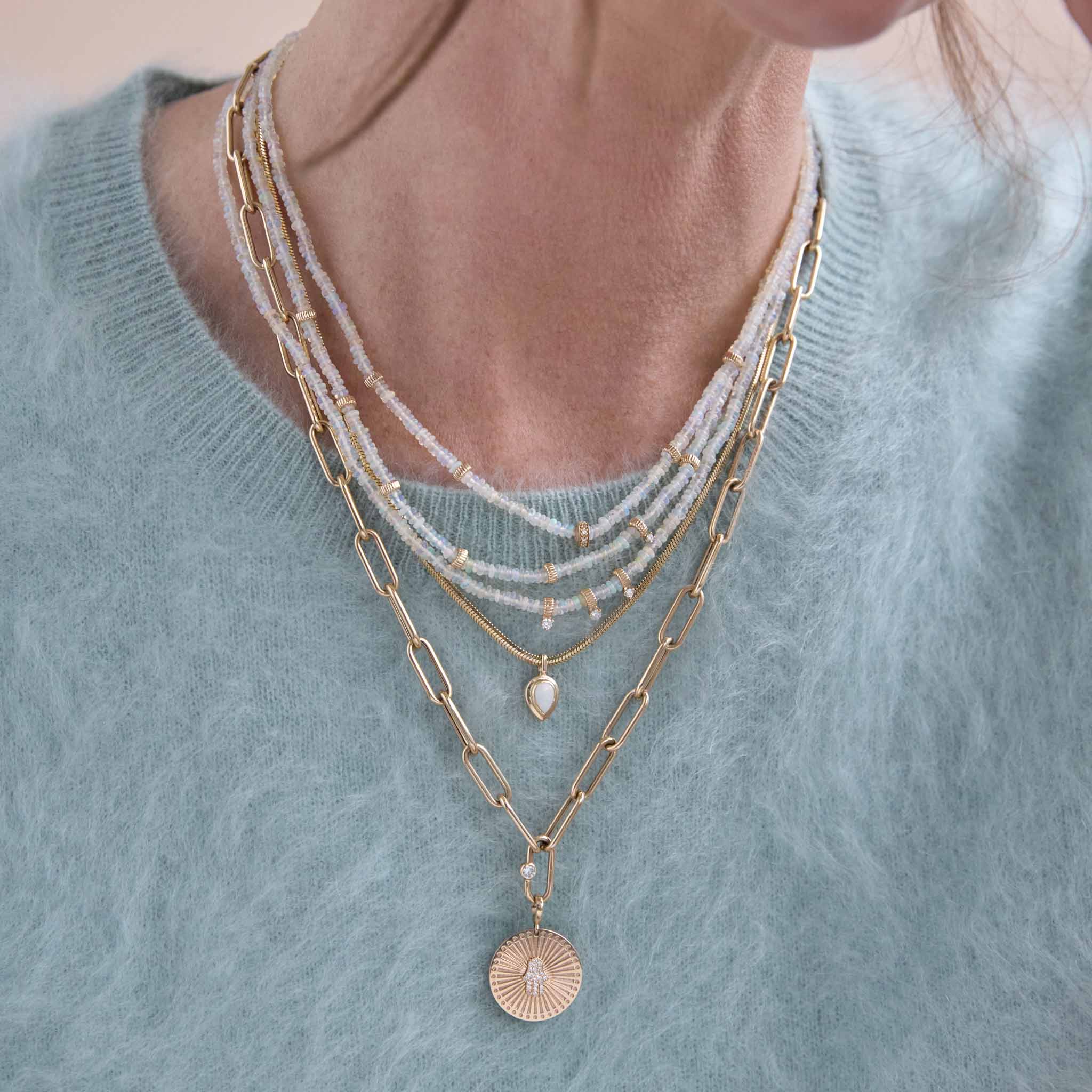 woman in a blue fuzzy sweater wearing three fire opal gemstone necklaces layered together with a Zoë Chicco 14k Gold Pear Opal Bezel Pendant Snake Chain Necklace and a 14k Gold Single Diamond Bezel Large Paperclip Chain Necklace with a Hamsa Sunbeam Medallion Pendant