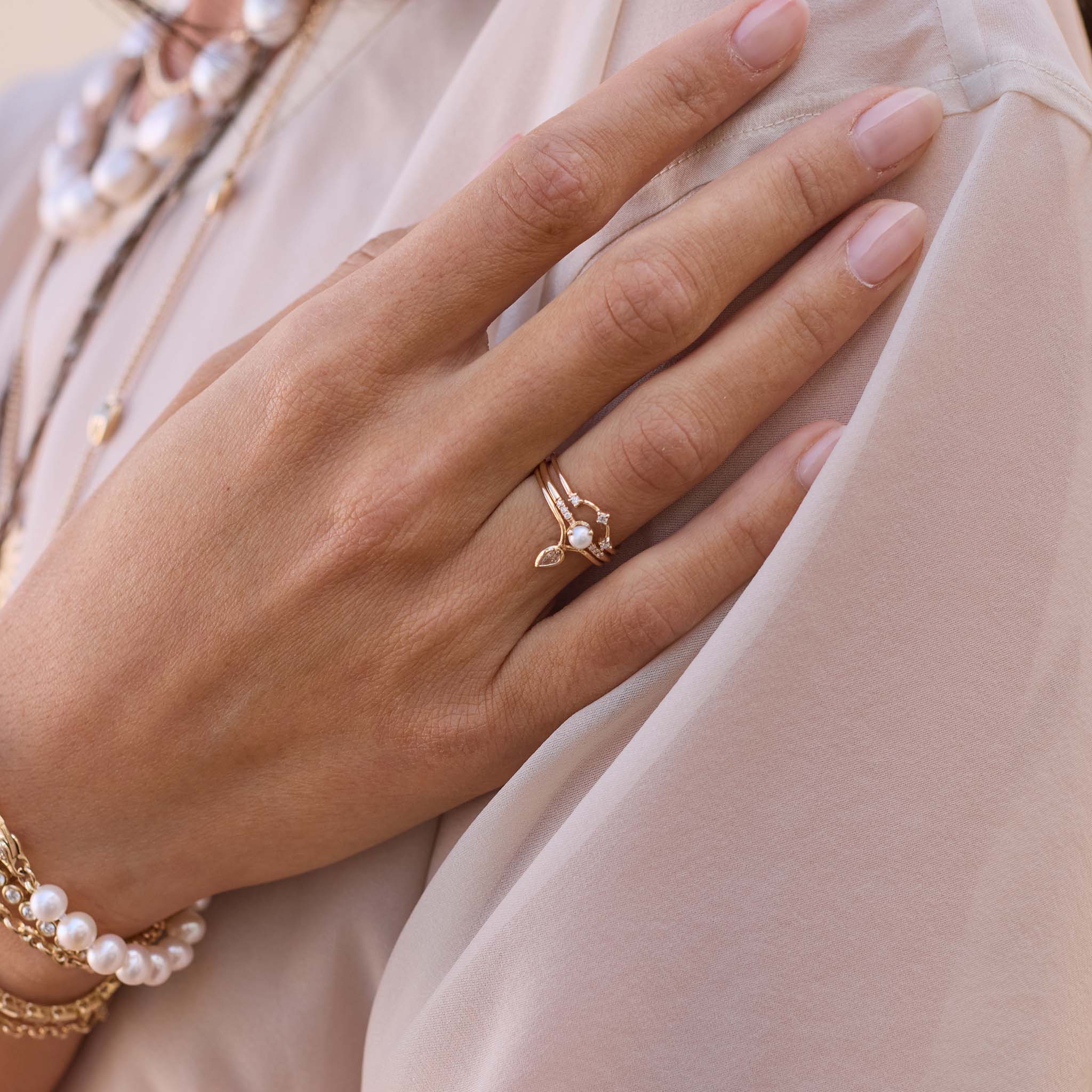 Close-up of a hand wearing gold pear and prong diamond ring and a pearl ring in the middle on a beige background