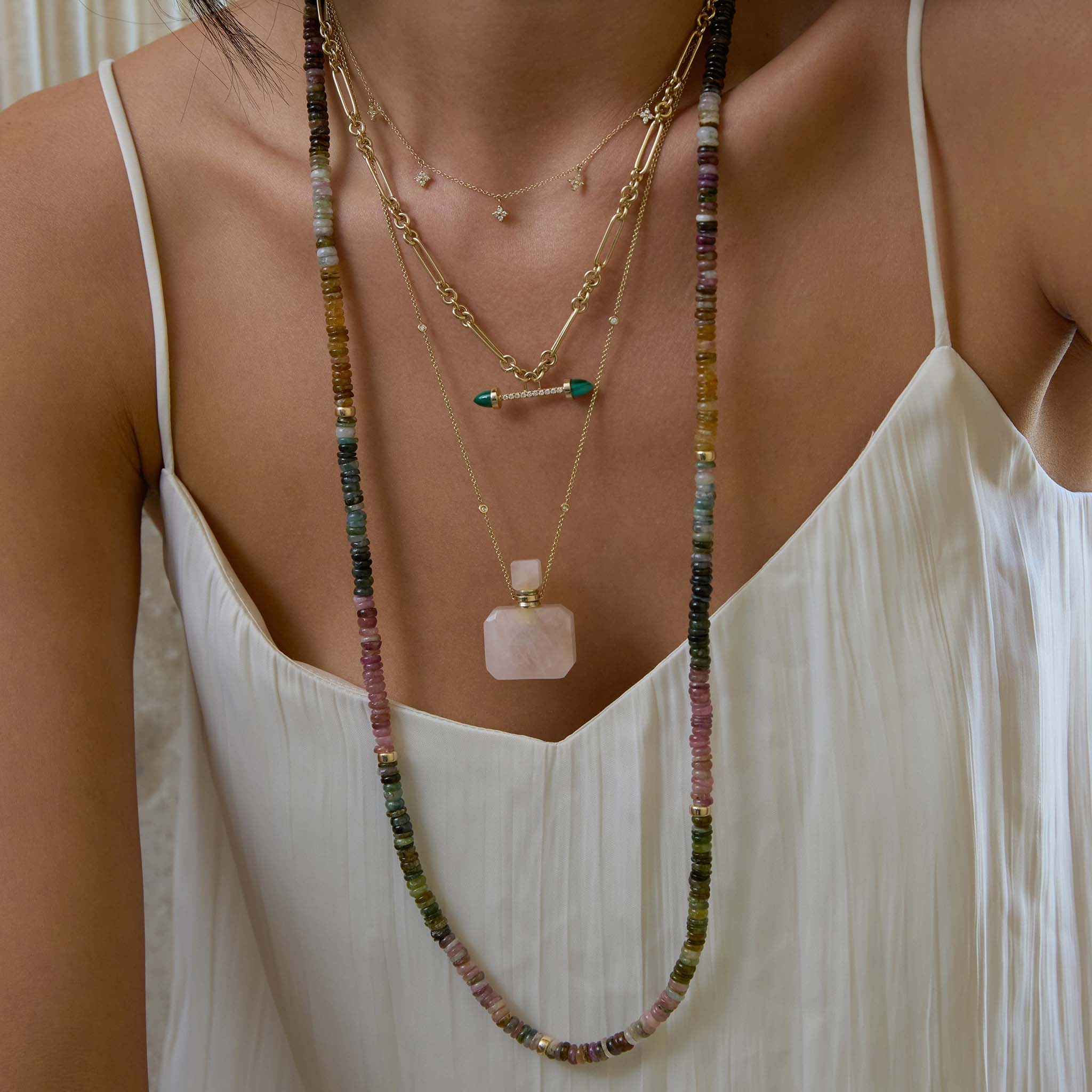 Layered Zoe Chicco 14k necklaces with diamonds and tourmaline gemstones with a rose quartz perfume bottle on a white textured shirt