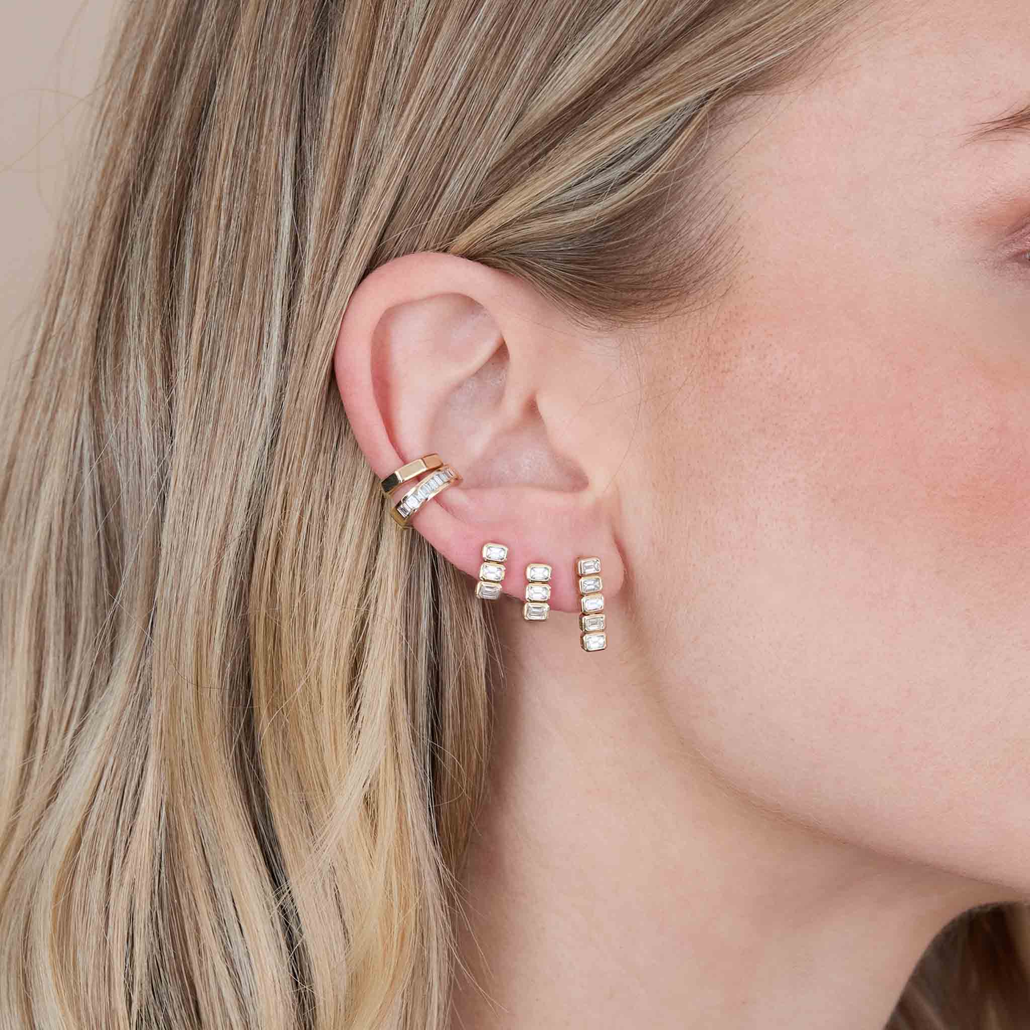 close up of a woman's ear wearing a Zoë Chicco 14k Gold 3 Horizontal Emerald Cut Diamond Tennis Drop Earring in both her second and third piercing and a 5 Horizontal Emerald Cut Diamond Tennis Drop Earring layered with a 14k Gold Octagon Ear Cuff and a Channel set Baguette Diamond Ear Cuff
