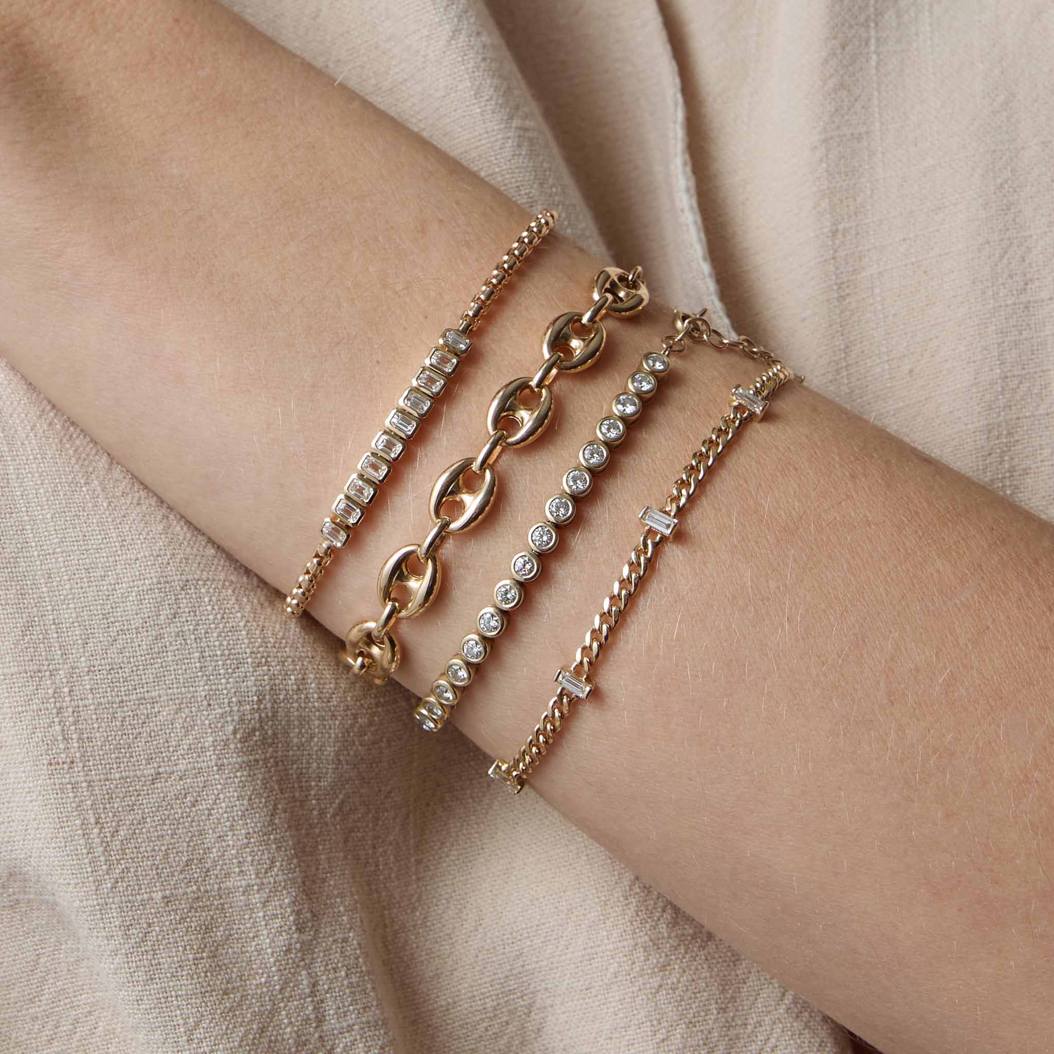 woman's wrist wearing a Zoë Chicco 14k Gold 2.5mm Diamond Bezel Tennis Bracelet stacked with a 14k 5 Vertical Baguette Diamond Station Small Curb Chain Bracelet, 14k Gold Large Puffed Mariner Chain Bracelet, and a 14k Vertical Emerald Cut Diamond Tennis Segment Box Chain Bracelet