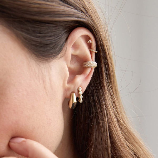 close up of a woman's ear wearing a Zoë Chicco 14k Gold Diamond Bezel Tennis Short Drop Earring in her second piercing and a 14k Gold Medium Wide Bamboo Hoop Earring in her first layered with a Pave Diamond Chubby Ear Cuff and a small prong set diamond trio stud