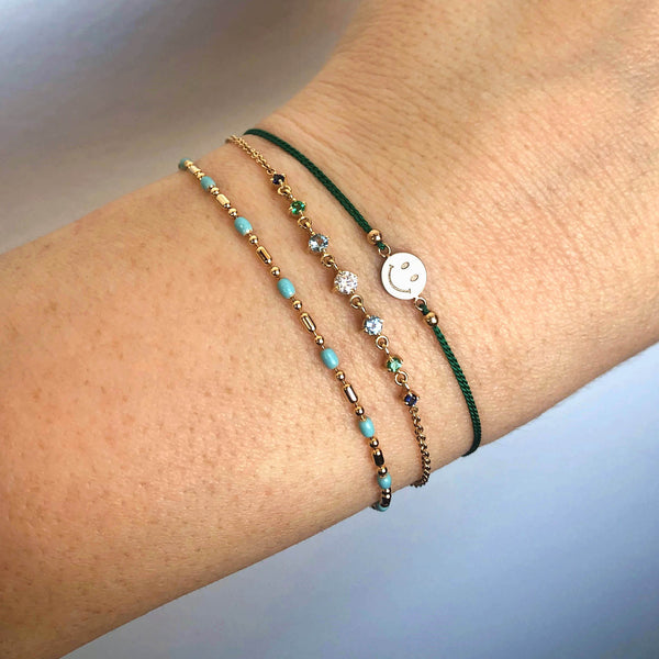 woman's wrist wearing a Zoë Chicco 14k Gold Midi Bitty Smiley Face Green Cord Bracelet stacked with a Linked Graduated Blue Ombre Gemstone Bolo Bracelet and an Turquoise Enamel Tube Bead Bracelet
