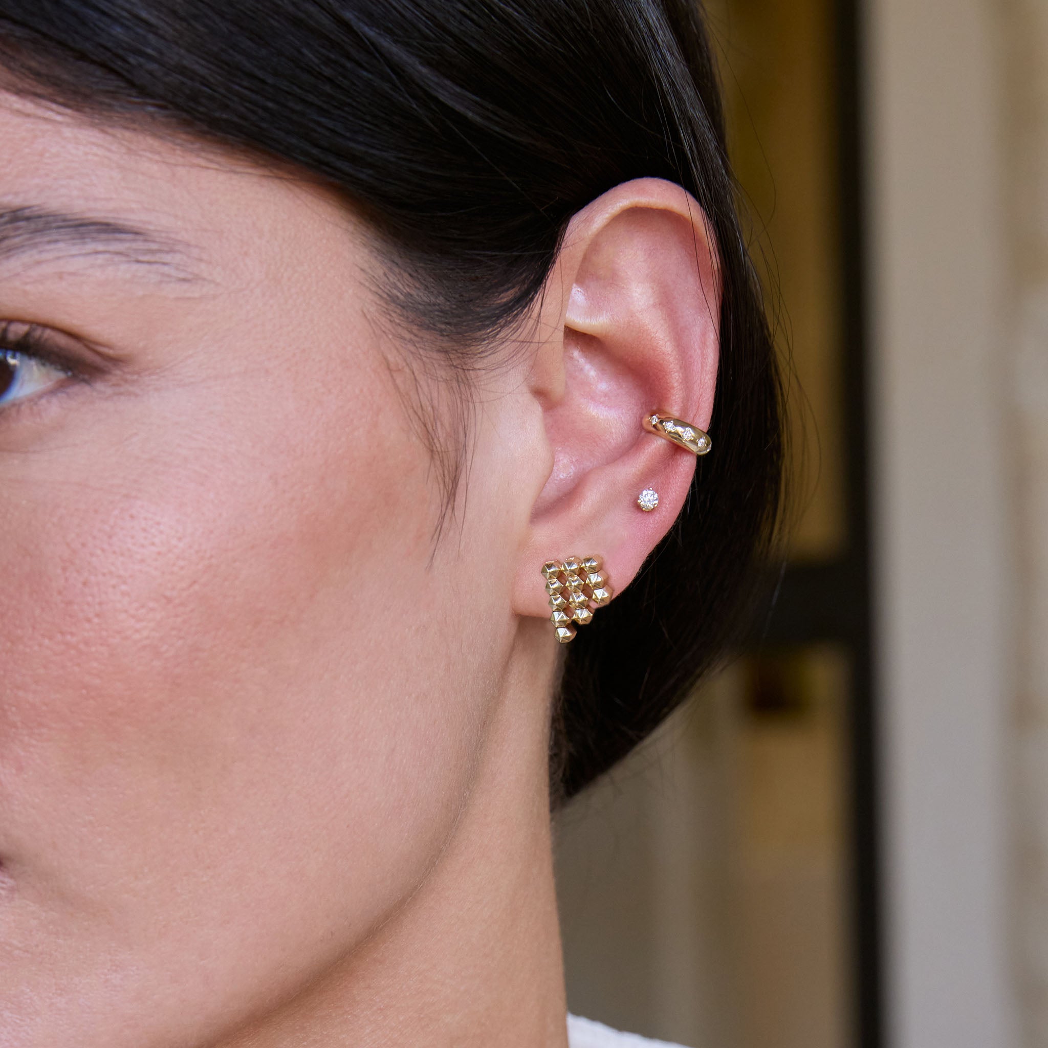 Close-up of a woman wearing Zoe Chicco 14k gold star stet diamond ear cuff, stud earring, and hexagon chain link waterfall earring on a blurred background