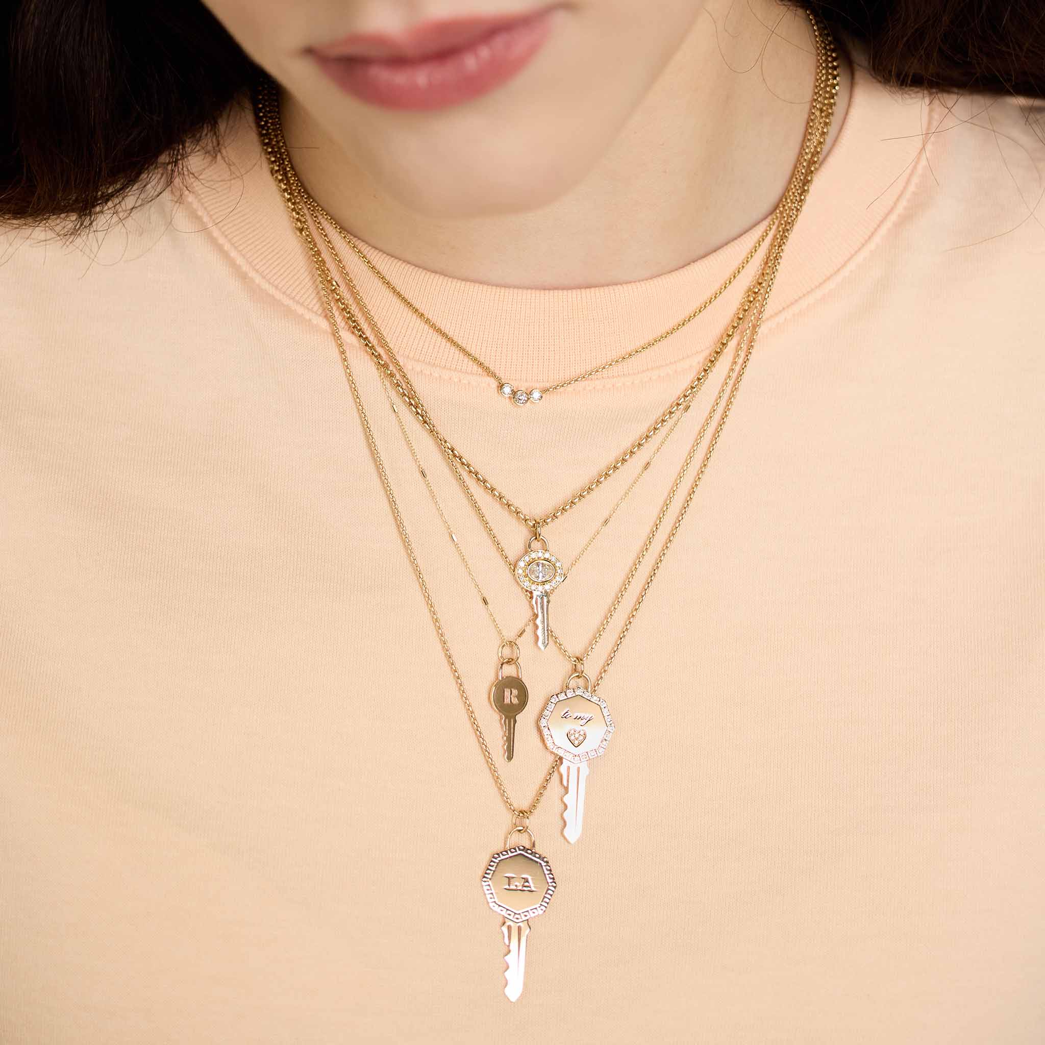 woman in a peach colored shirt wearing a Zoë Chicco 14k Gold Engraved Initial Letter "R" Key Pendant Necklace layered with an 14k Oval Diamond Halo Key Necklace, 14k Pavé Diamond Key to my Heart Pendant Box Chain Necklace, and a 14k Single Los Angeles "LA" Key Charm Pendant on a box chain