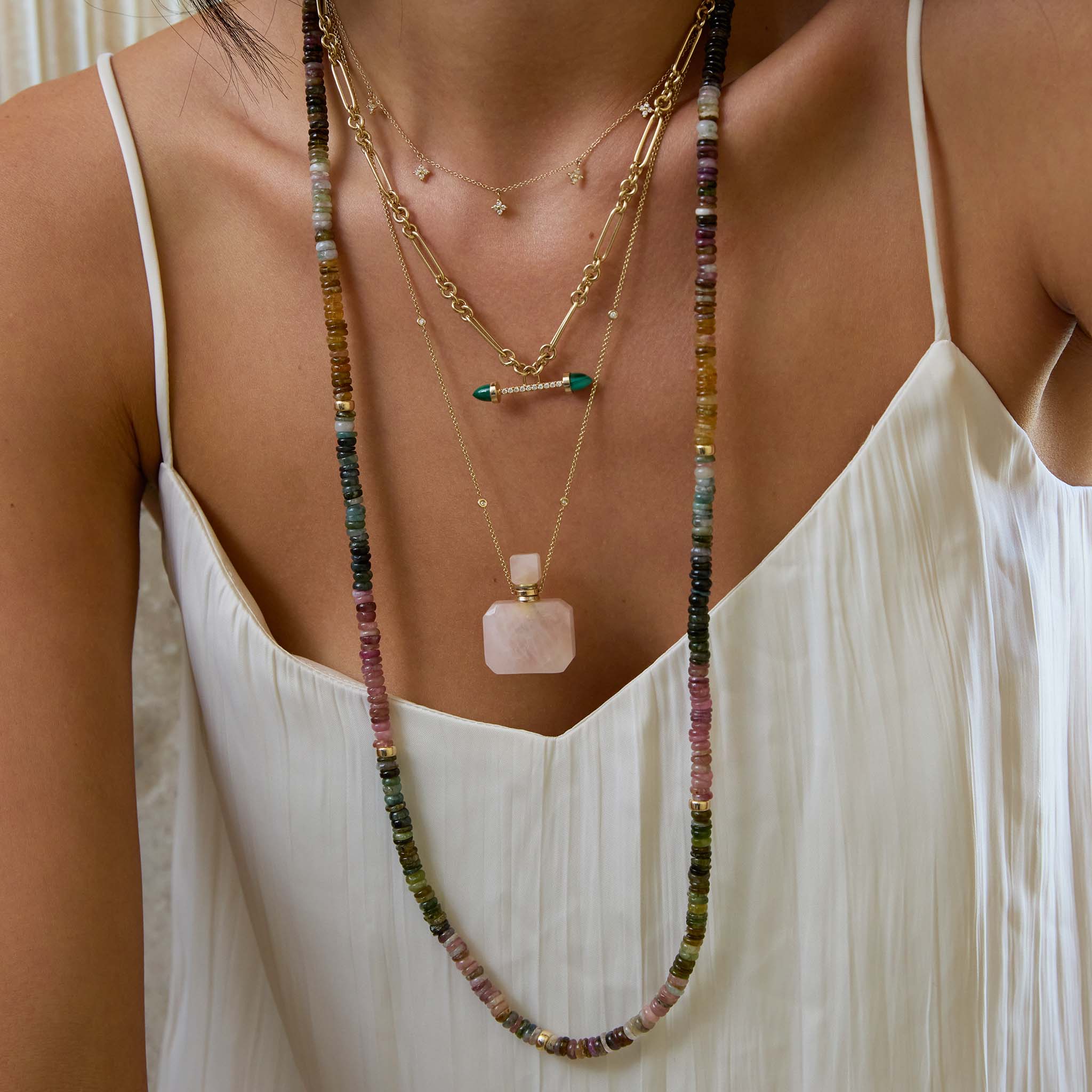 Close-up of a woman wearing a white blouse and multiple Zoe Chicco colored gemstone bead necklaces with 14k gold and diamonds and a square rose quartz perfume bottle chain necklace