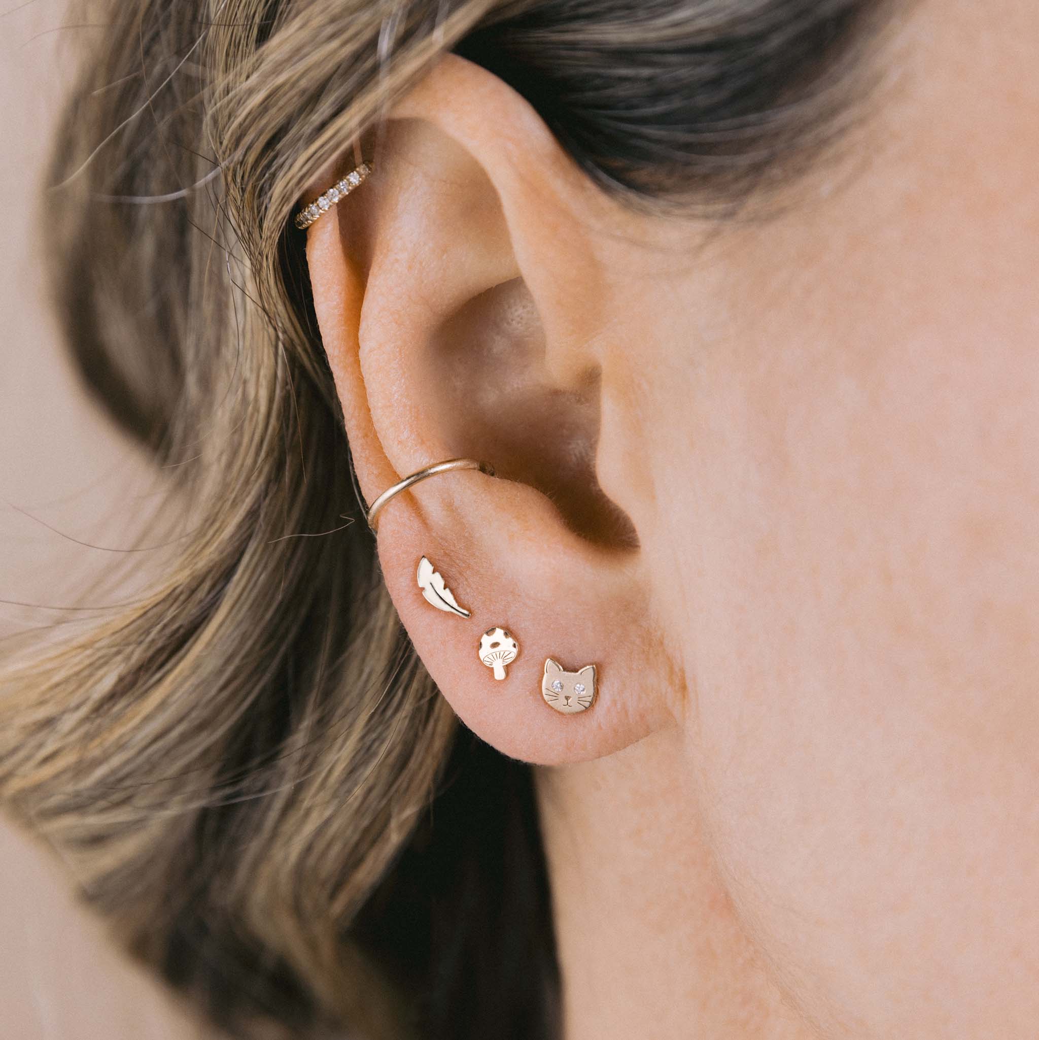 A woman wears Zoe Chicco's 14k Itty Bitty Feather Stud, Mushroom Stud and Cat stud with diamond eyes