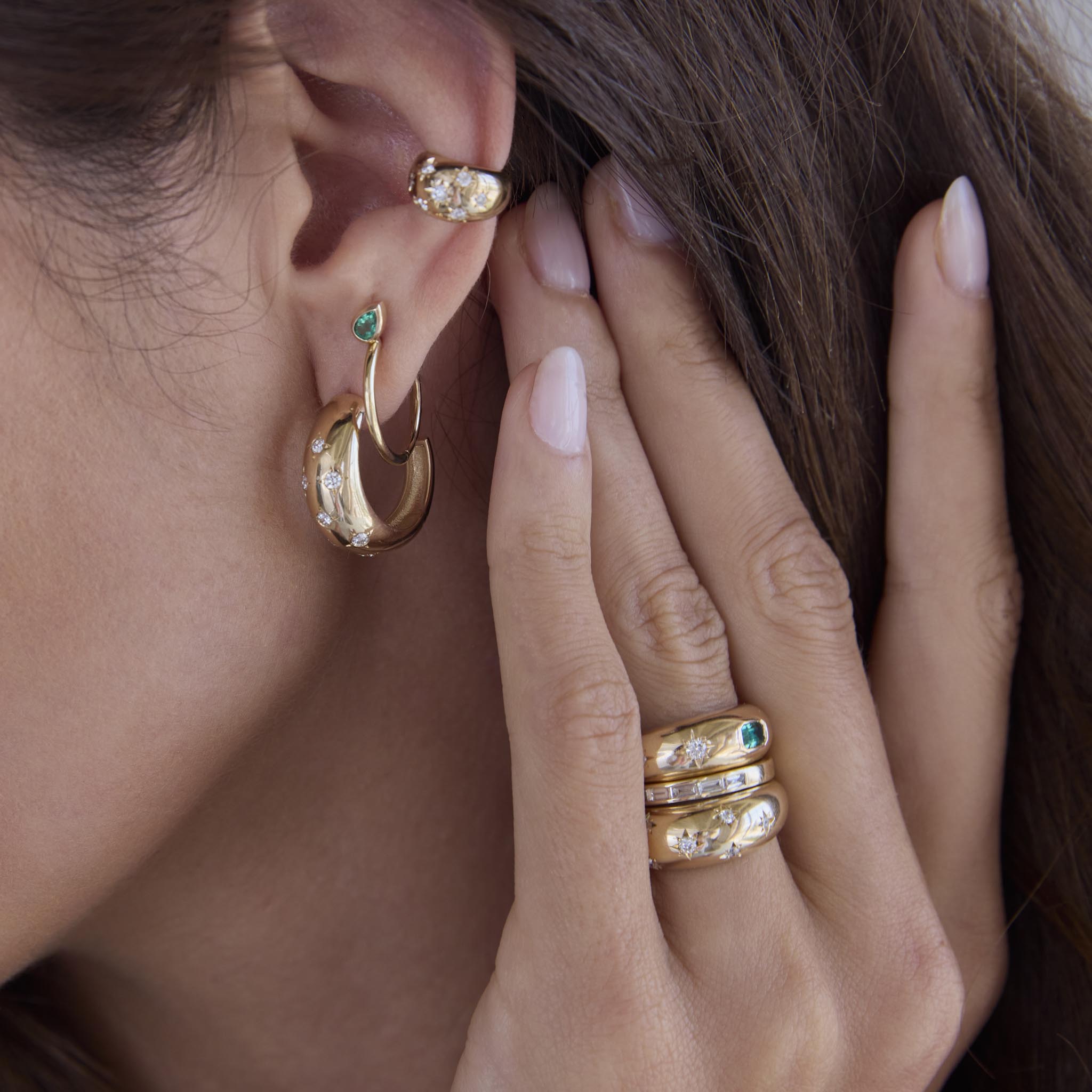 woman with her hand raised next to her ear wearing a Zoë Chicco 14k Gold Emerald Cut Emerald & Graduating Diamonds Small Aura Ring stacked with two diamond rings and wearing a Scattered Diamond Medium Aura Hoop and a Pear Emerald Huggie Hoop in her ear