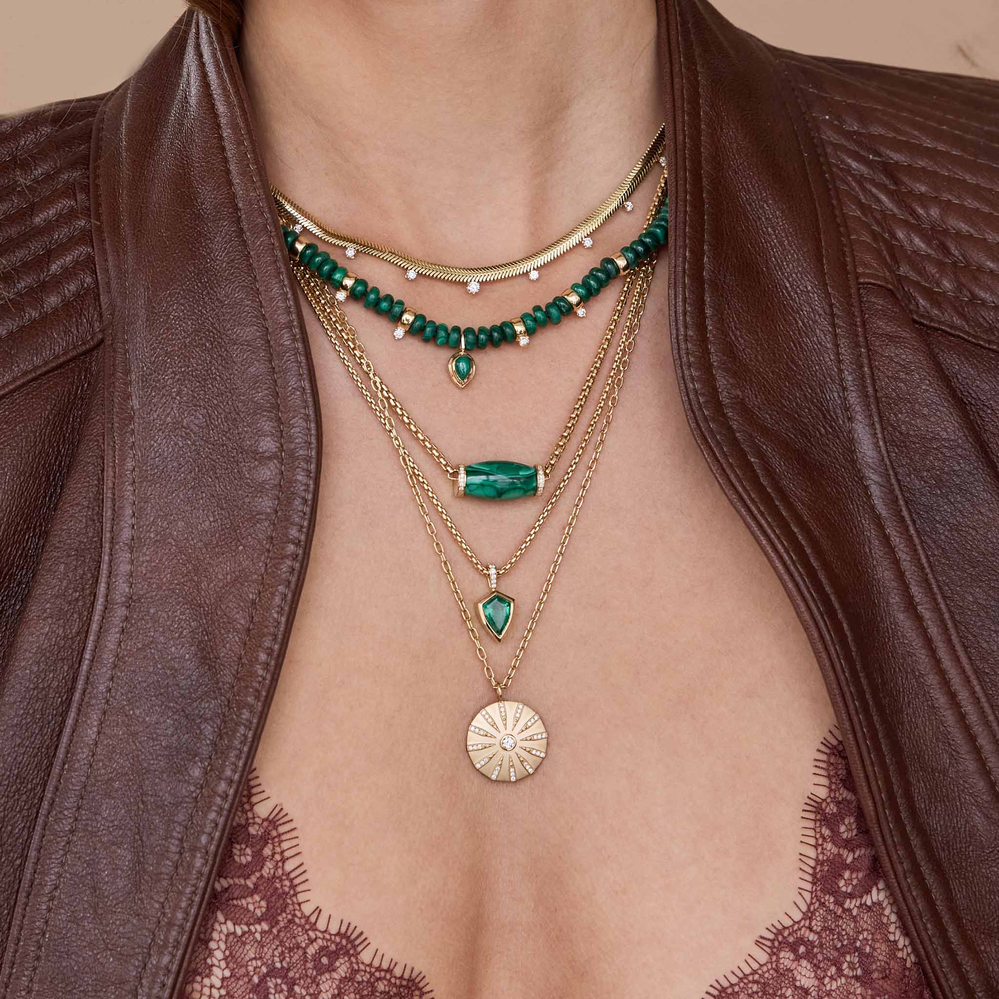Close-up of layered necklaces 14k Diamond Malachite Barrel Bead Medium Box Chain Necklace, emerald and malachite necklaces, and gold pendants on a person wearing a brown leather