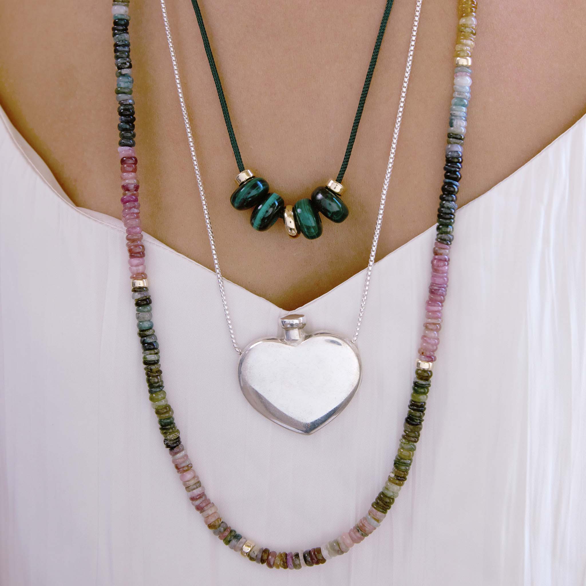 Close up of a neck with layered Zoe Chicco gemstone bead necklaces and a sterling silver repurposed Tiffany & Co heart shaped perfume bottle against a white shirt