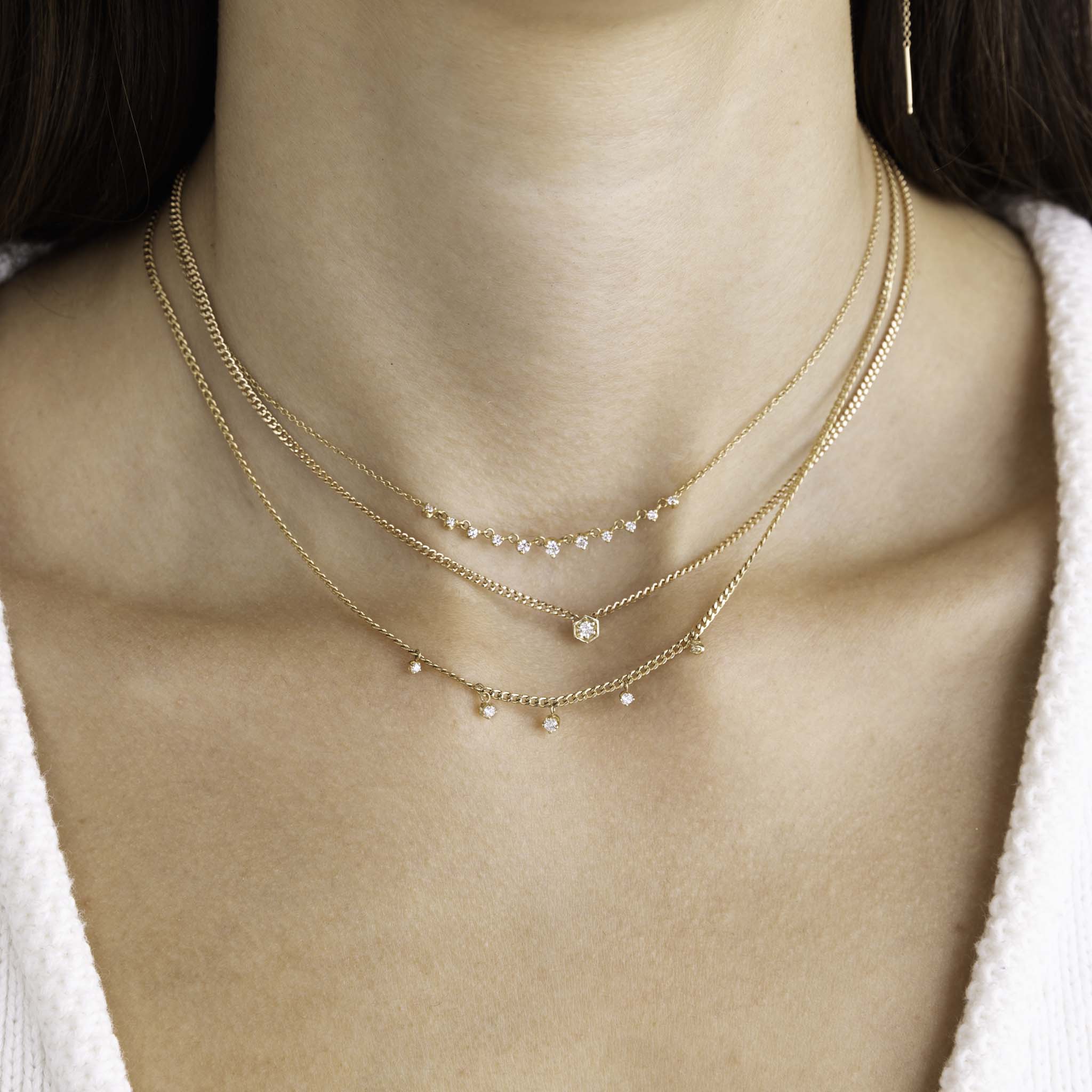 A woman with dark hair is wearing a Zoe Chicco necklace stack including 14k 9 Linked Prong Diamond Necklace, 14k Gold Diamond Hexagon Curb Chain Necklace, and 14k 5 Dangling Prong Diamond Extra Small Curb Chain Necklace