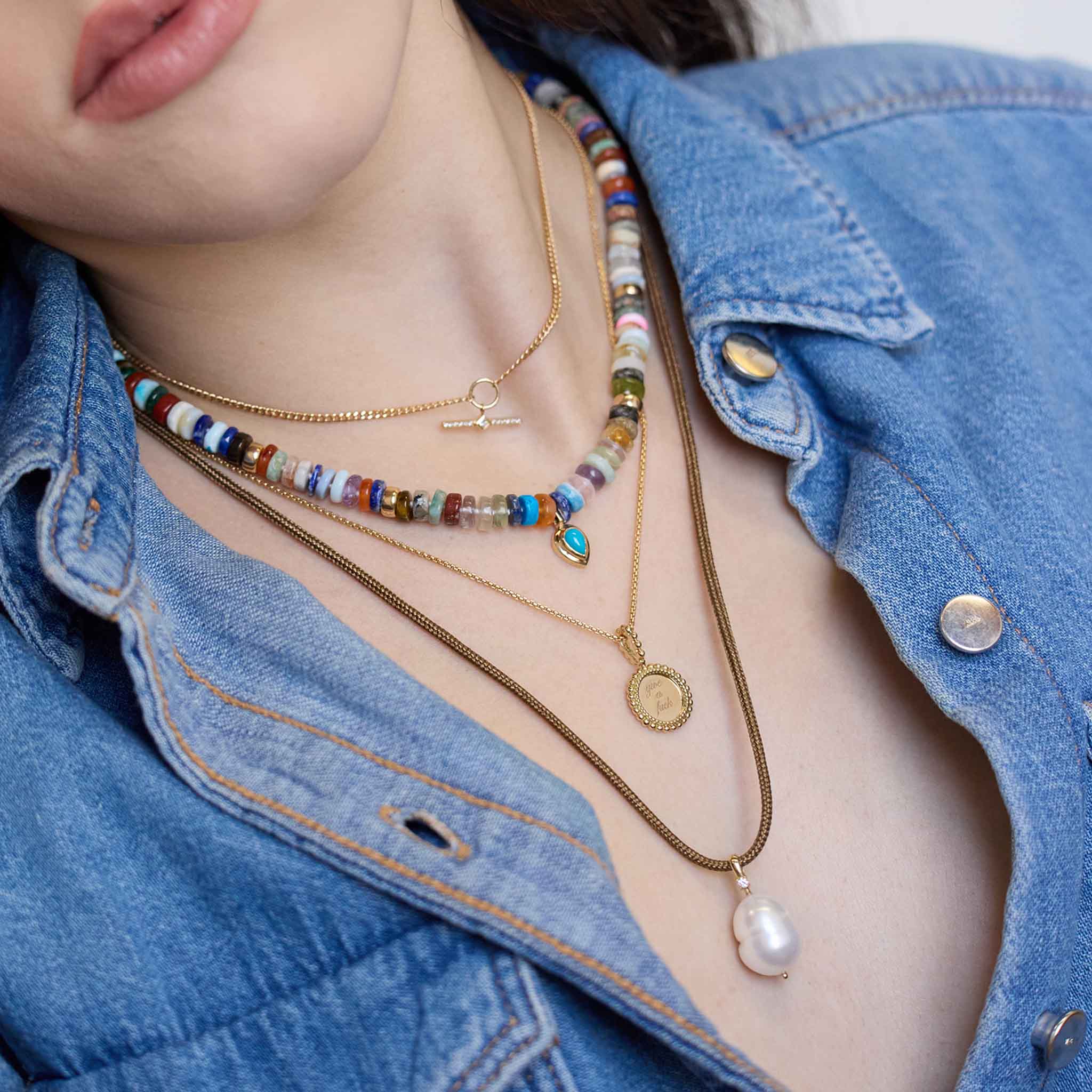 woman in a denim shirt wearing a Zoë Chicco 14k Gold "give a fuck" Beaded Disc Pendant Box Chain Necklace layered with a 14k Mixed Dark Gemstone Rondelle Bead Necklace with Pear Turquoise Pendant and a 14k Baroque Pearl & Diamond Bail Pendant Paracord Necklace
