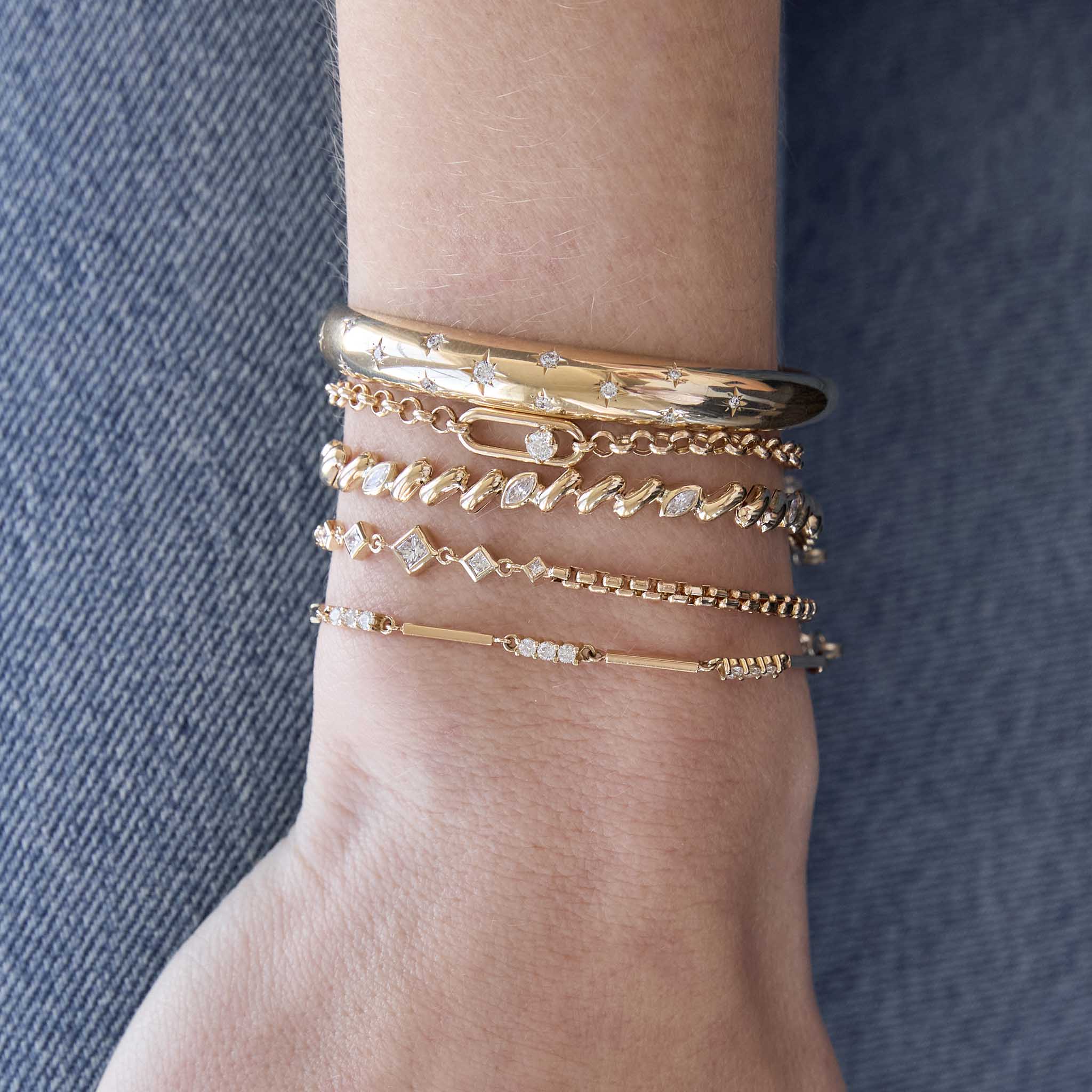 woman's wrist against a denim background wearing a Zoë Chicco 14k Gold Single Nested Diamond Large Paperclip Link Medium Rolo Bracelet layered with a 14k Scattered Star Set Diamonds Small Aura Cuff, 14k 5 Marquise Diamond Station San Marco Chain Bracelet, 14k Linked Graduated Princess Diamond Bezel Box Chain Bracelet, and a 14k Gold and Diamond Linked Bar Bracelet