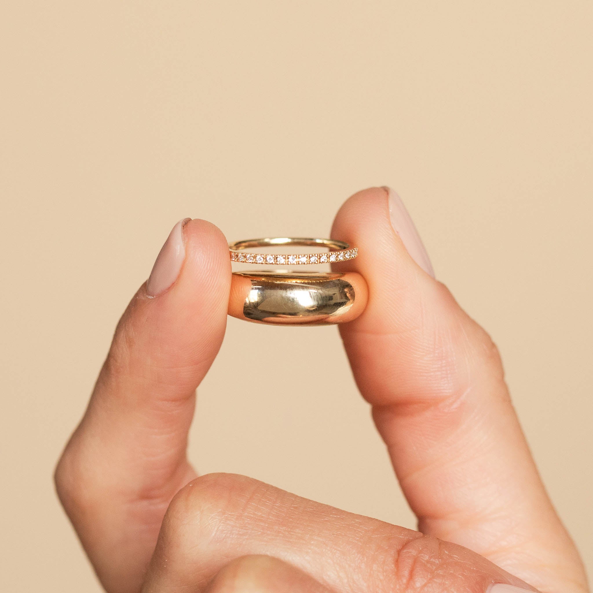 Against a beige background is a woman holding 2 rings between her fingers: a single plain Aura ring and our half pave diamond eternity band.