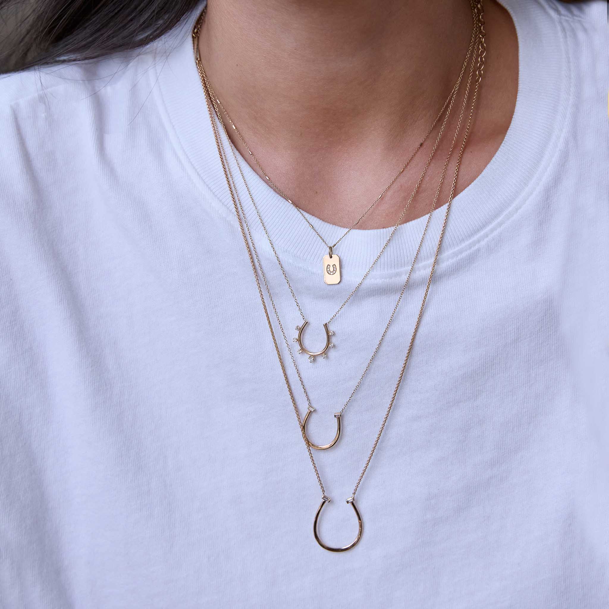 Close-up of a person wearing multiple Zoe Chicco 14k gold and diamond horseshoe shaped pendant necklaces on a white shirt