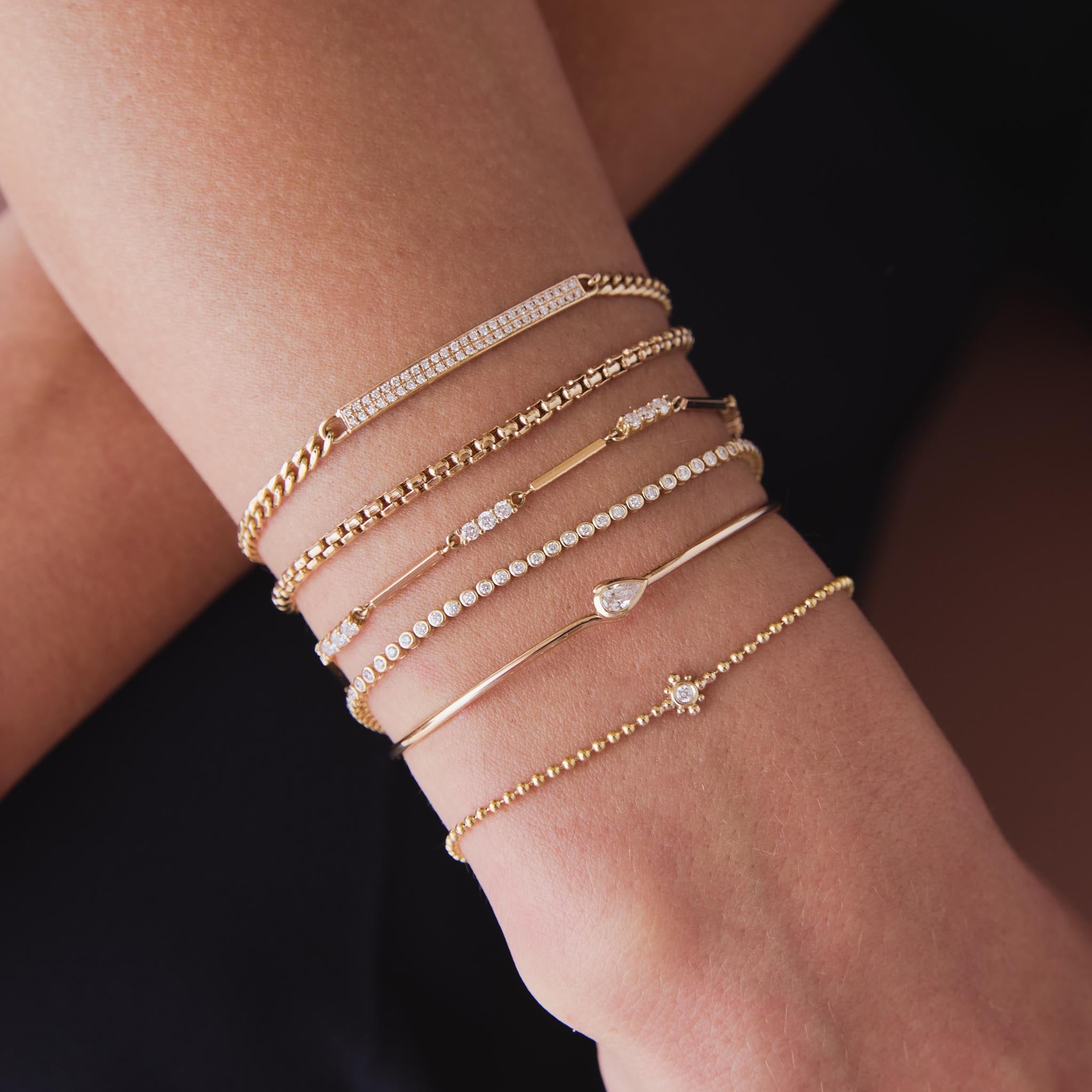 woman's wrist wearing a Zoë Chicco 14k Gold Pear Diamond Bezel Wire Cuff Bangle Bracelet stacked with a Diamond Bezel Tennis Bracelet, a Medium Box Chain Bracelet, a Pavé Diamond ID Bar Curb Chain bracelet, and a 14k Mixed Gold & Diamond Bar Bracelet