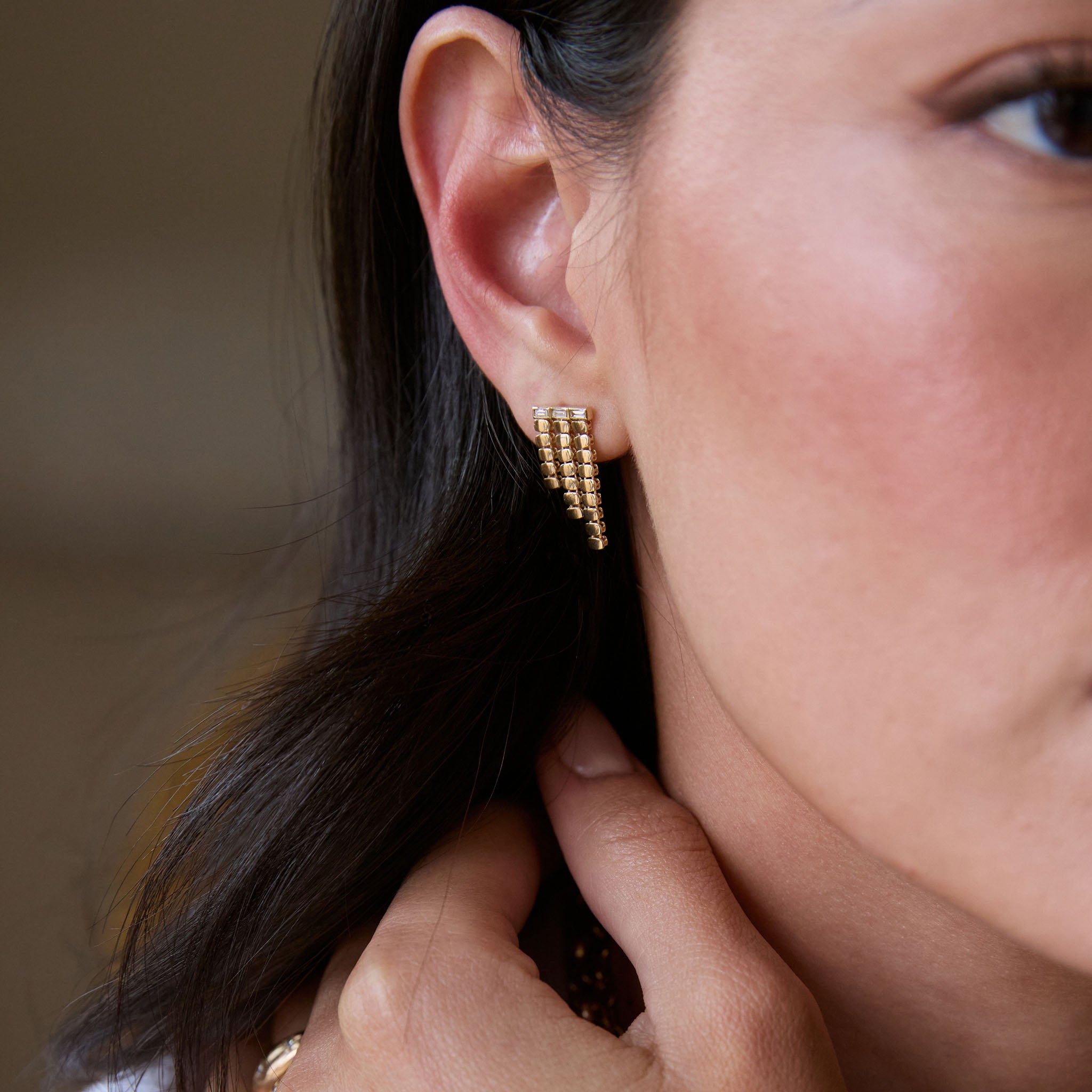 Close-up of a woman wearing a Zoe Chicco 14k gold oval chain waterfall stud earring with baguette diamonds against a neutral blurred background