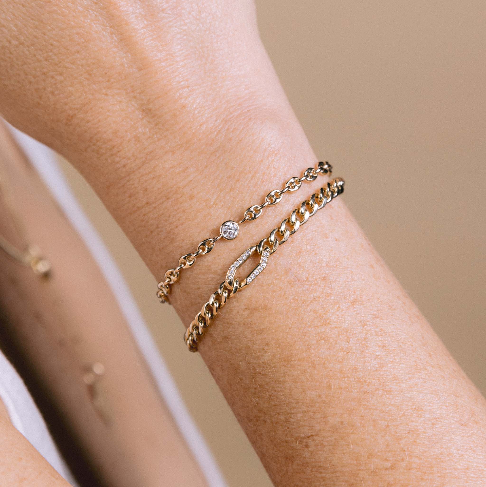 A woman is wearing the Zoë Chicco 14k Gold Floating Diamond Small Puffed Mariner Chain Bracelet and a 14k Gold Pavé Diamond Oval Link Medium Curb Chain Bracelet stacked together on her wrist
