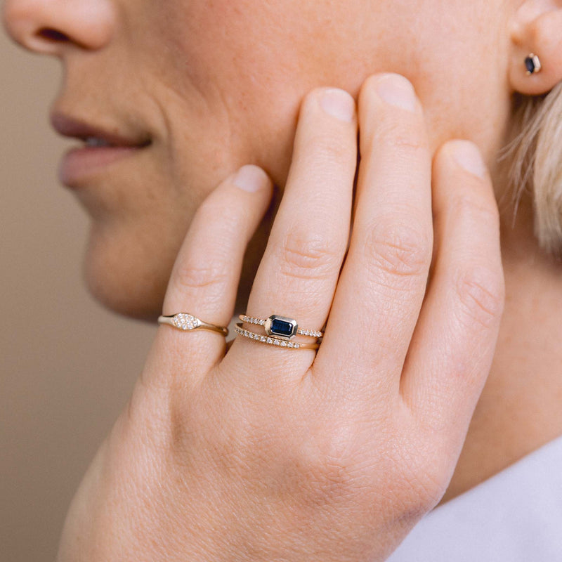 woman resting her hand on her cheek wearing a Zoë Chicco 14k Gold Emerald Cut Blue Sapphire Bezel Pavé Diamond Band Ring stacked with a Pavé Diamond Half Eternity Band and a Pavé Diamond Oval Signet Ring on her pinky finger