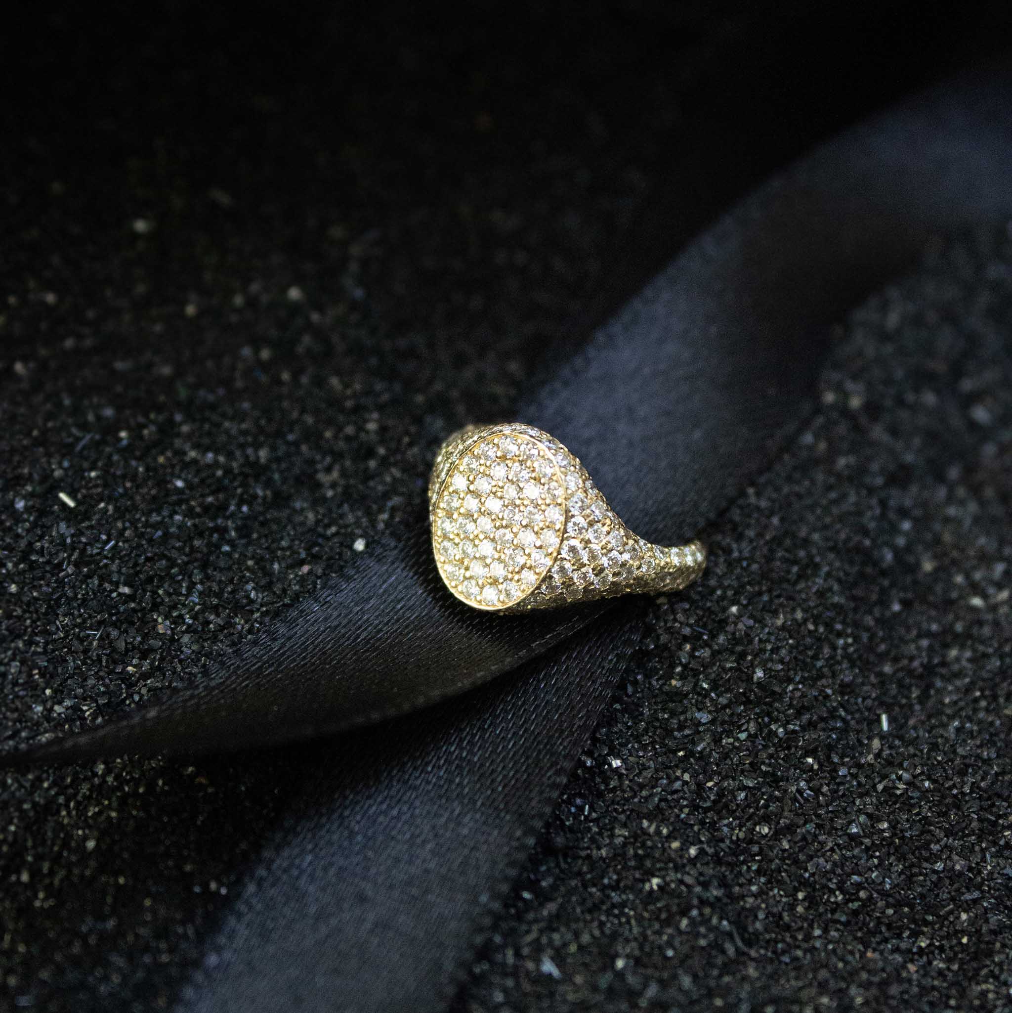 A pave diamond encrusted signet ring is sitting on black ribbon and black sand. The ring has an oval face and is often styled as a pinky ring.