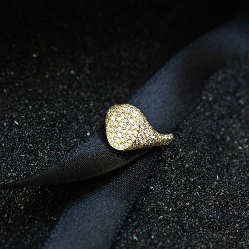 A pave diamond encrusted signet ring is sitting on black ribbon and black sand.  The ring has an oval face and is often styled as a pinky ring.