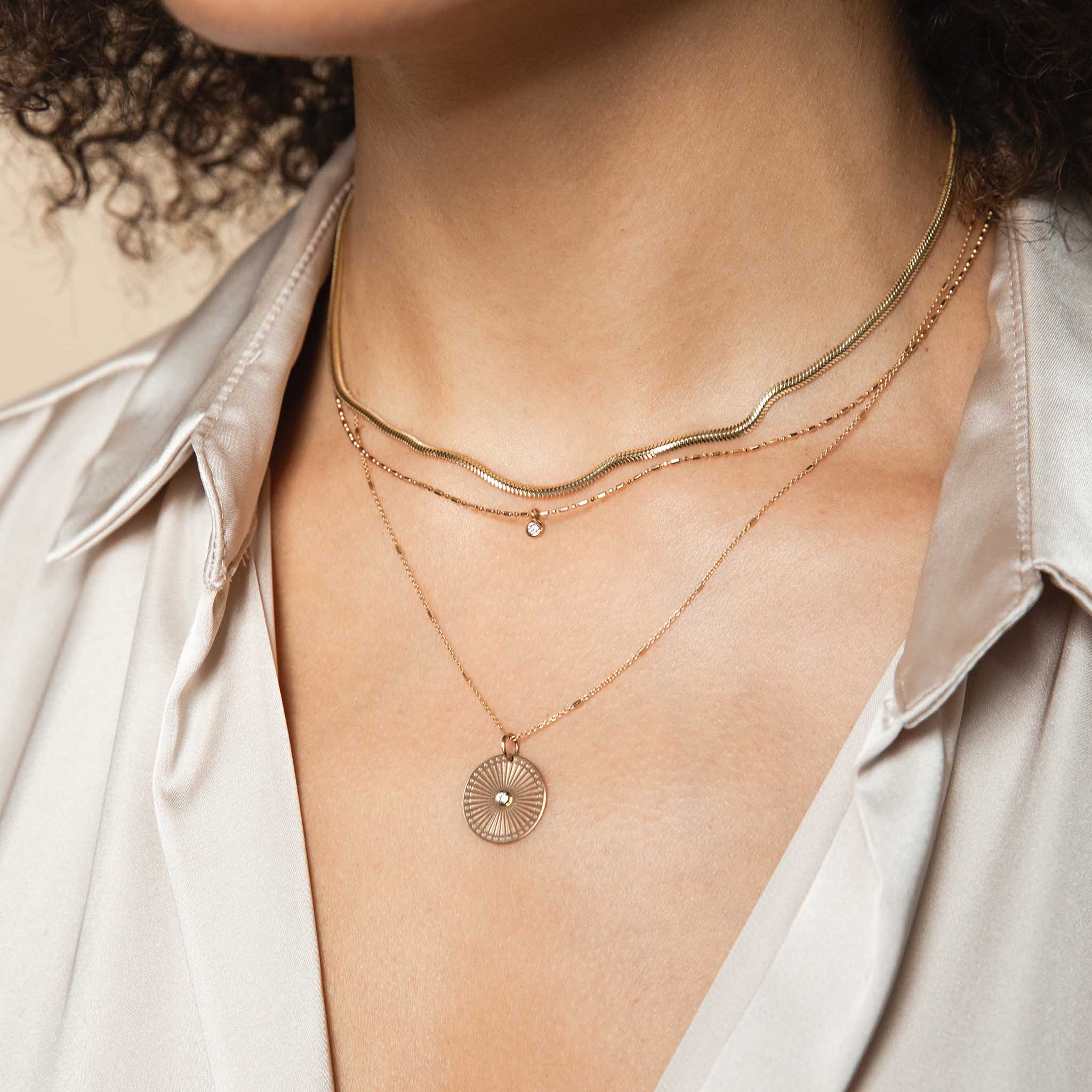 A woman wearing a silk shirt has 3 layered necklaces around her neck: the small snake chain, simple solitaire diamond and our classic Small Sunbeam Medallion Necklace