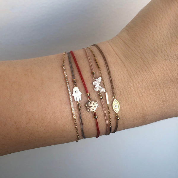 close up of a woman's wrist against a white background wearing four different Zoë Chicco 14k Gold Midi Bitty Cord Bracelets (hamsa, ladybug, butterfly, evil eye) stacked together with two bar bracelets
