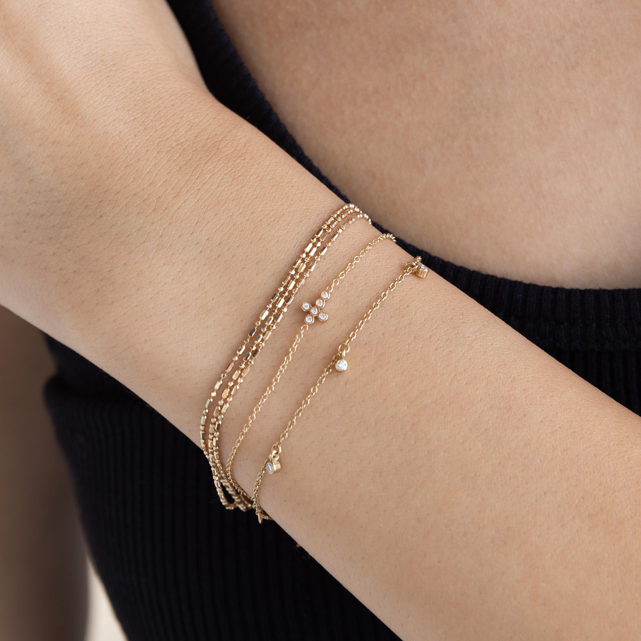 close up of a woman's wrist wearing a Zoë Chicco 14k Gold Small Horizontal Diamond Bezel Cross Bracelet stacked with a Triple Wrap Tube Bead Chain Bracelet and a Dangling Diamond Bezel Station Bracelet