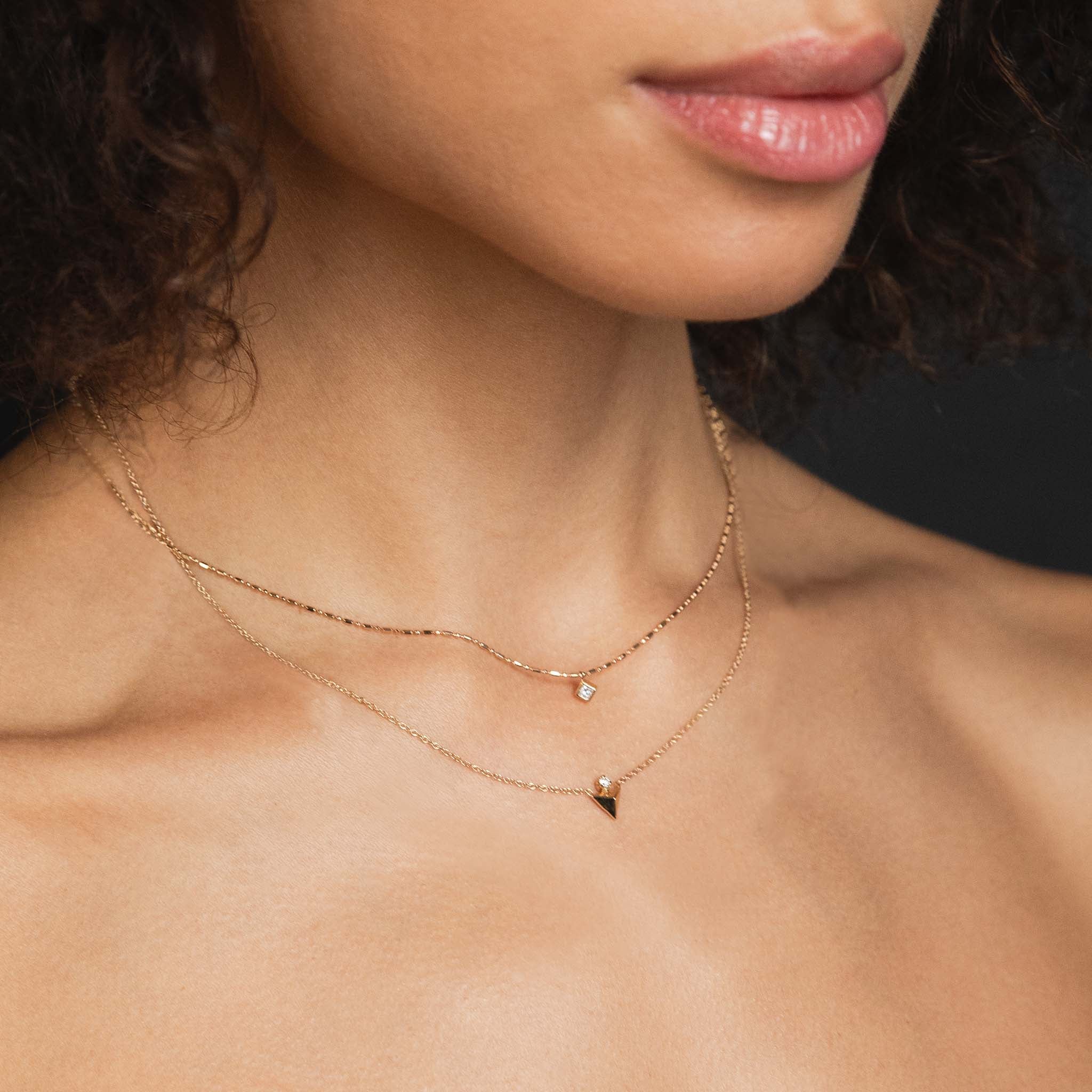 A woman wears two necklaces: small princess diamond drop and a pyramid necklace with prong diamond.