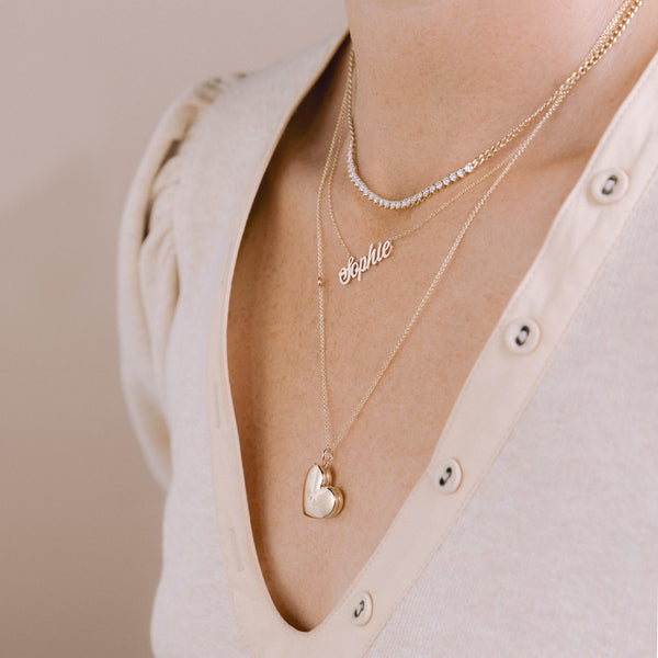 woman in a beige top wearing a Zoë Chicco 14k Gold 2 Initial Letters Heart Locket Necklace engraved with an R and M layered with a Custom Script Letter Necklace with the name Sophie and a Prong Diamond Tennis Segment Necklace