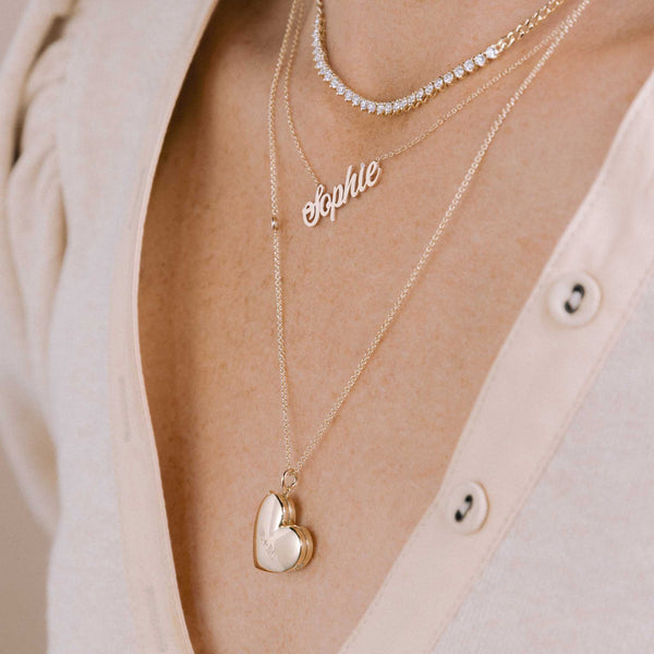 A woman in a pale pink top wearing two Zoë Chicco personalized 14k gold necklaces layered together. Top necklace reads Sophie in script letters and bottom is a heart locket engraved with R+M