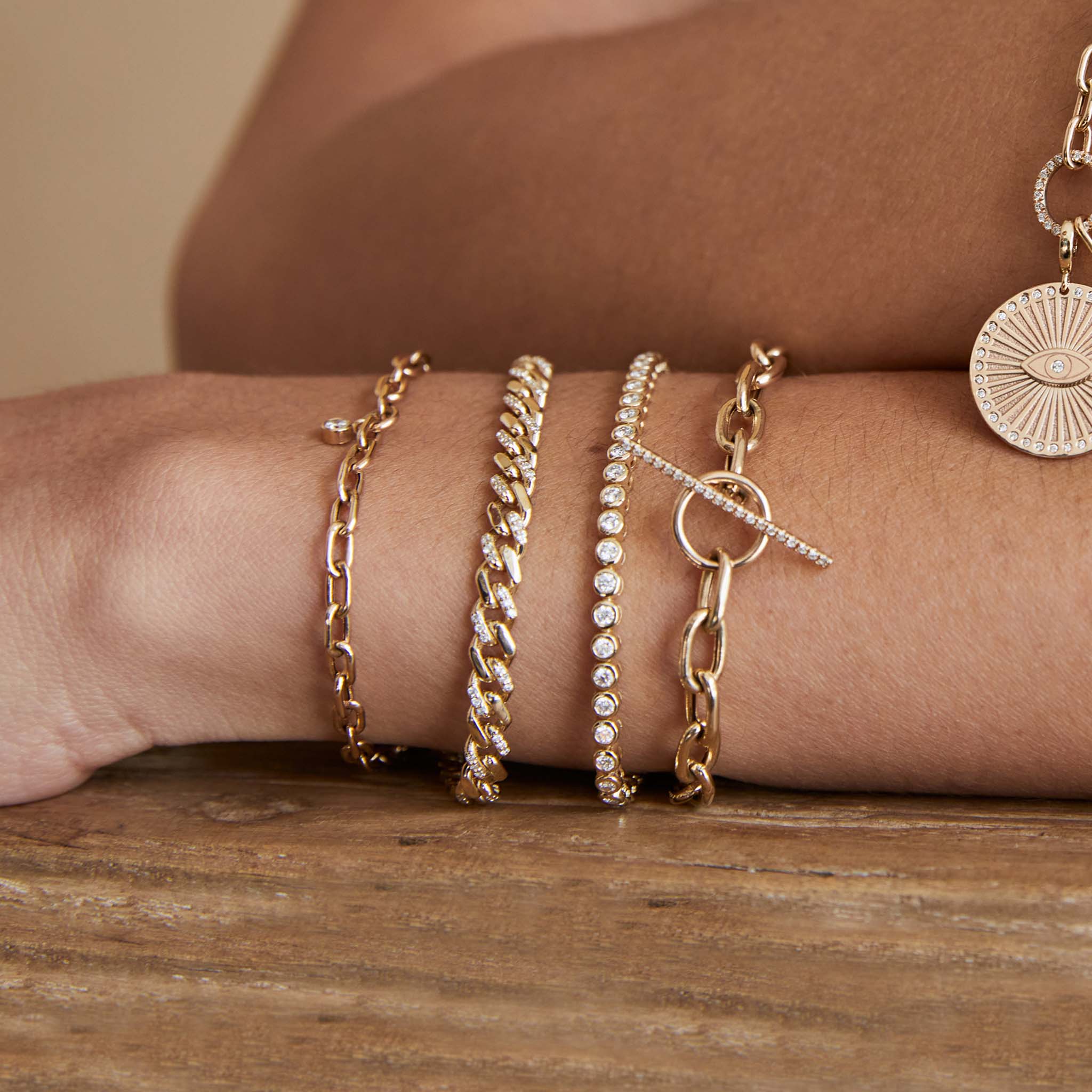 close up of woman's wrist laying on a wood table wearing a Zoë Chicco 14k Gold 2mm Diamond Bezel Tennis Bracelet layered with three heavy chain bracelets