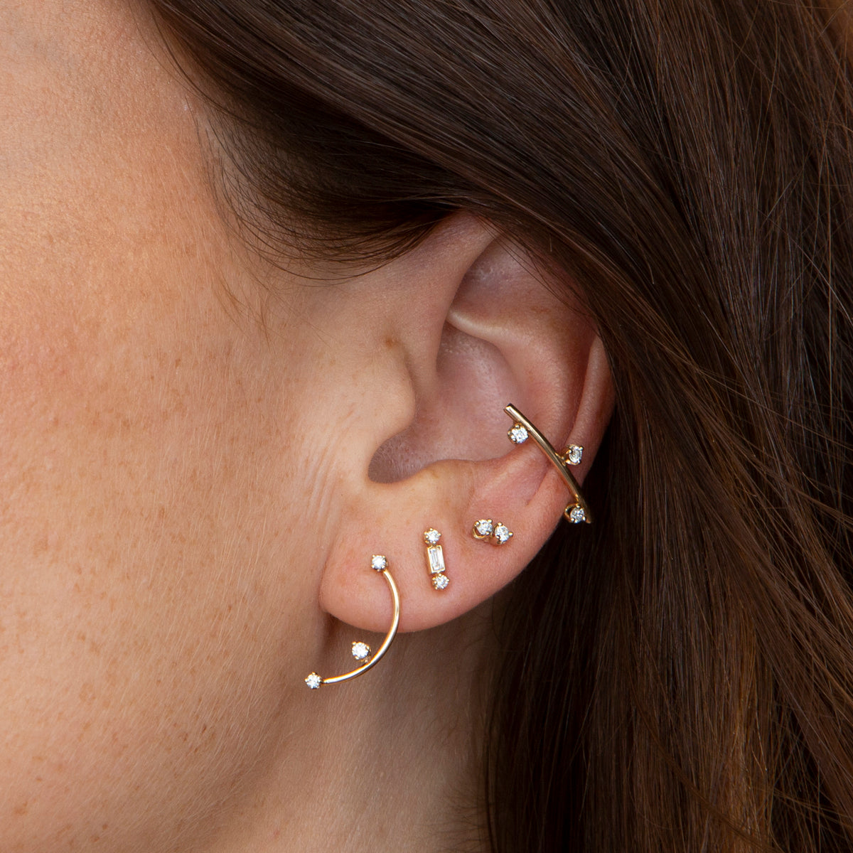 woman's ear wearing Zoë Chicco 14k Gold Twin Diamond Stud Earring, a Baguette & Prong Diamonds Stud, a 3 Prong Diamond Curved Bar Drop Earring, and a 3 Scattered Prong Diamonds Thick Wire Bar Ear Cuff