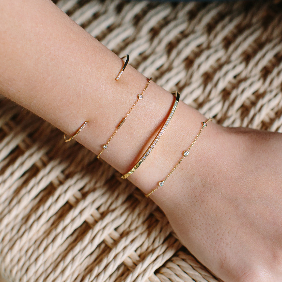 woman's wrist resting on a woven rattan chair wearing a Zoë Chicco 14k Gold Horizontal Bar & Floating Diamond Station Bracelet