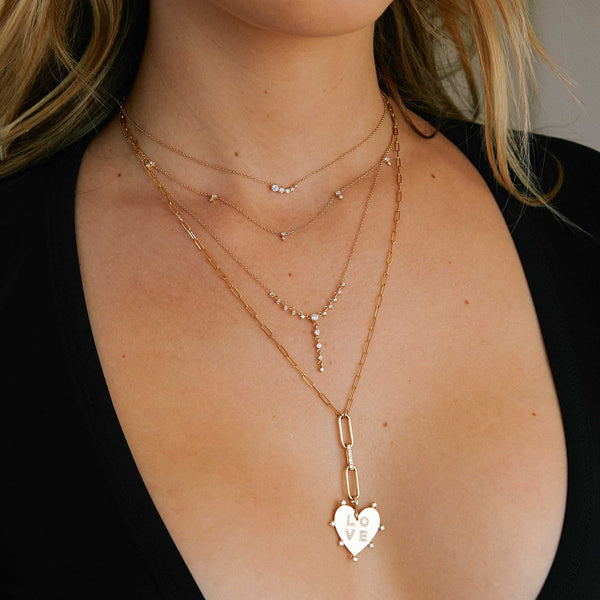 woman in a black top wearing a Zoë Chicco 14k Gold Mixed Paperclip Chain Lariat Necklace with Baguette Diamond Link with a 7 Prong Diamond Pave Diamond LOVE Heart Charm Pendant dangling from the bottom of the chain and layered with three other diamond necklaces