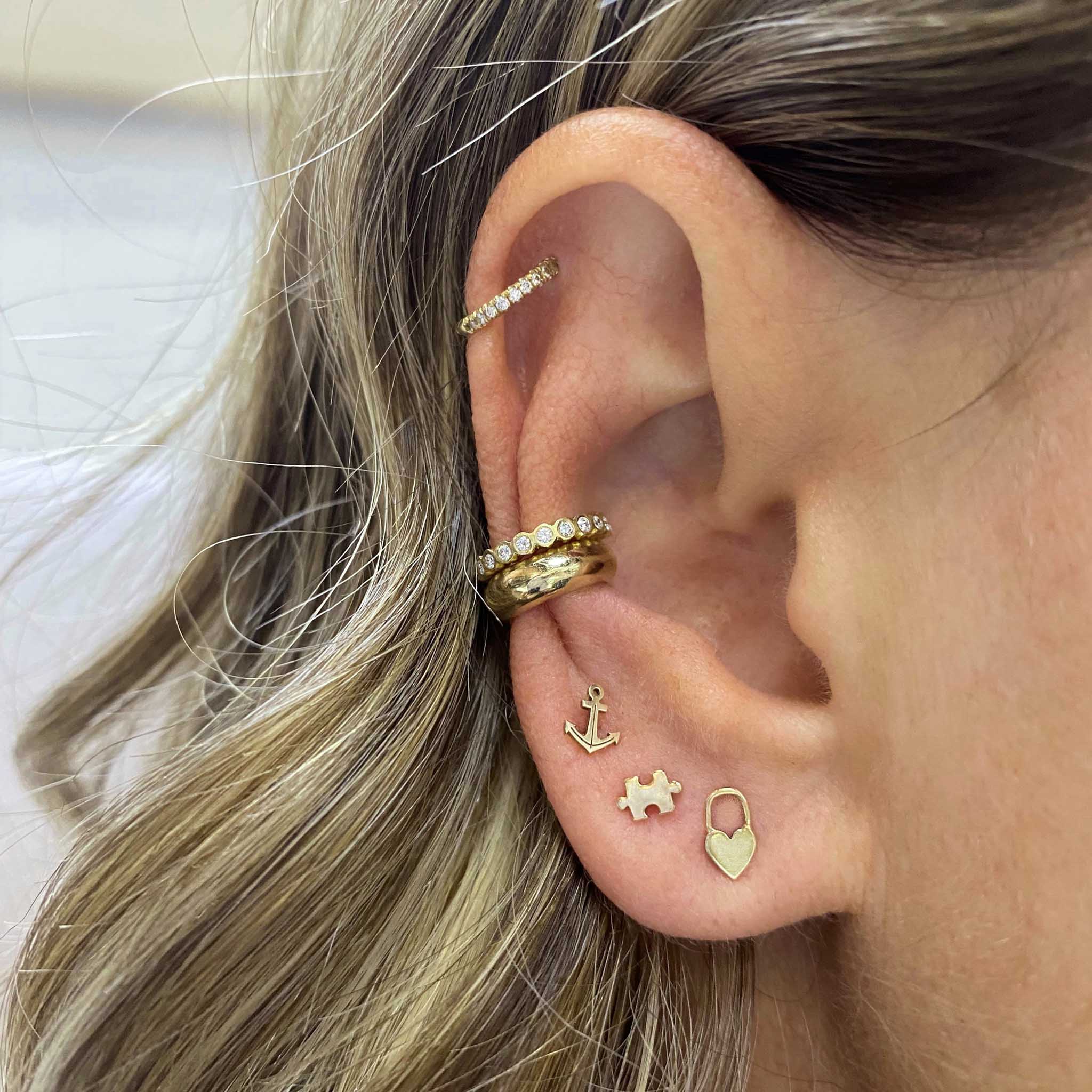 Woman's ear wearing Zoe Chicco 14k Gold Itty Bitty Heart Padlock Stud Earring with an Itty Bitty Puzzle Stud, Itty Bitty Anchor Stud, wide gold ear cuff and two diamond ear cuffs
