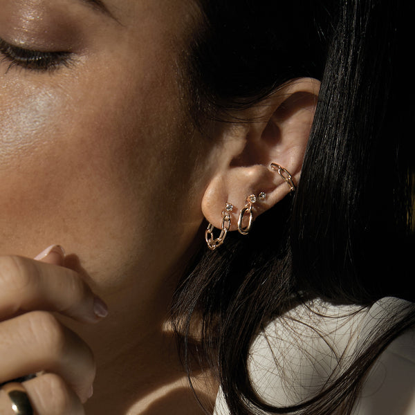 close up of woman with dark hair wearing a Zoë Chicco 14k Gold Solid Medium Square Oval Link Chain Ear Cuff layered with a Medium Prong Diamond Solitaire Stud, Prong Diamond with Single XL Square Oval Link Earring, and a Prong Diamond Medium Square Oval Link Huggie Hoop