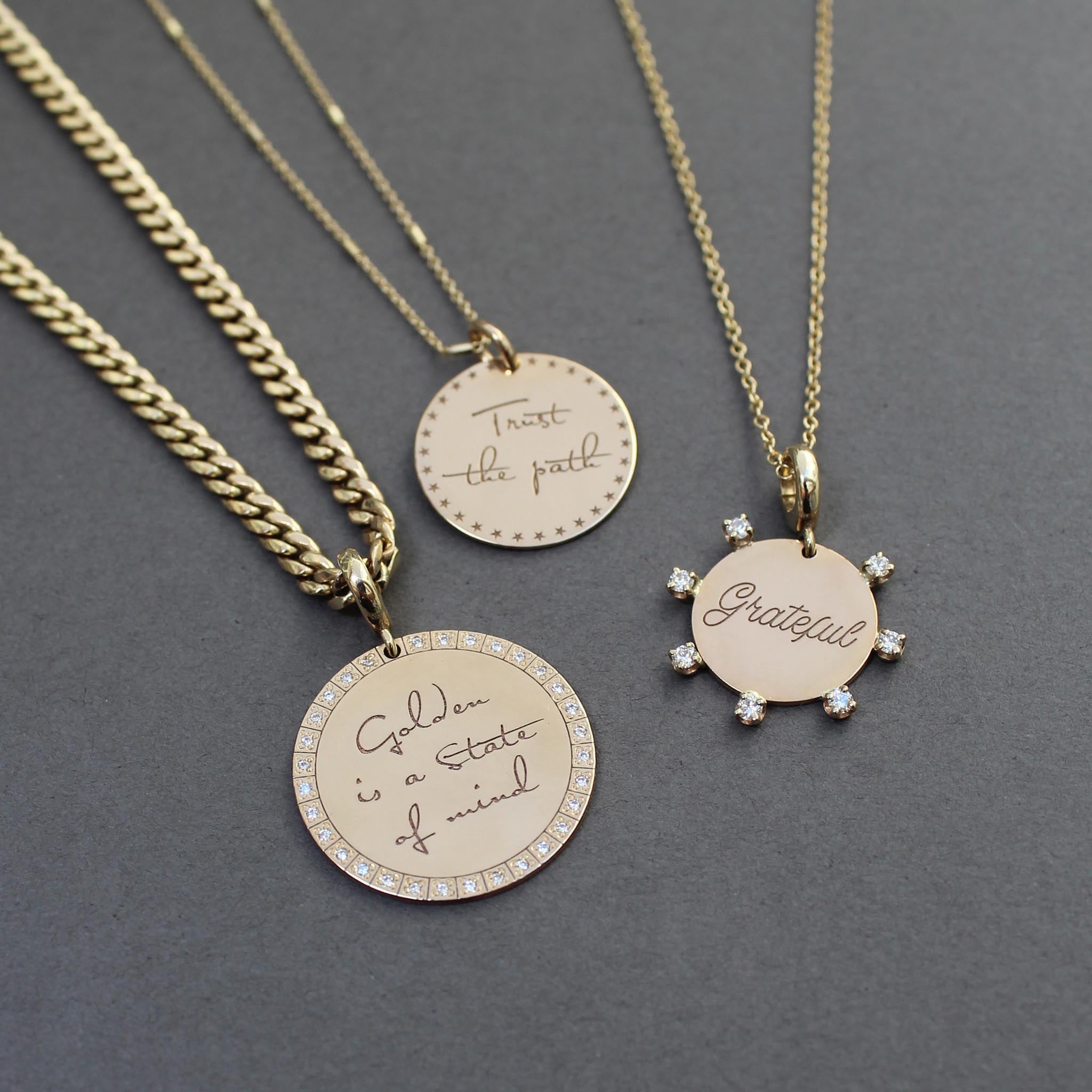 A Zoë Chicco 14k Gold Medium Mantra with Diamond Border Charm Pendant on a curb chain laying flat against a dark gray background