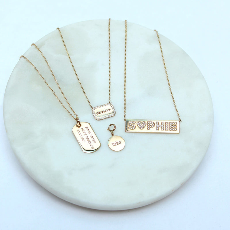 A Zoë Chicco 14k Personalized Rounded Rectangle Nameplate Necklace laid flat with other necklaces and a charm