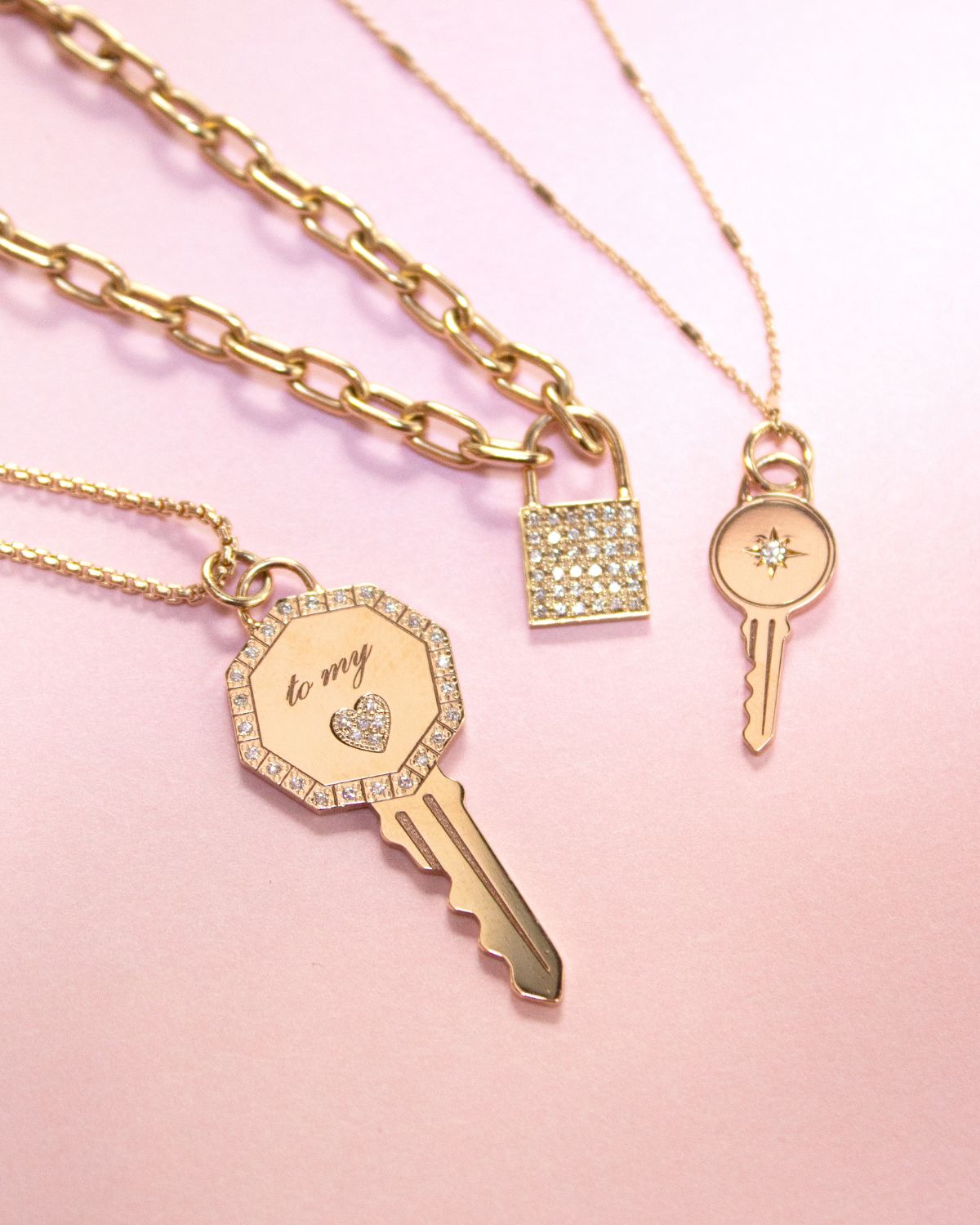Three Zoe Chicco 14k gold and diamond necklaces for the Tokens of Love Gift Guide laying against a pink background
