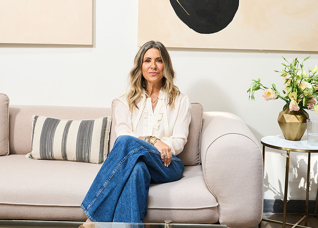 Woman sitting on a couch wearing dark jeans and a white top with a neutral background