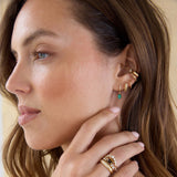 Close-up of a woman wearing multiple Zoe Chicco 14k gold earrings with diamonds and green stone and rings on a neutral background